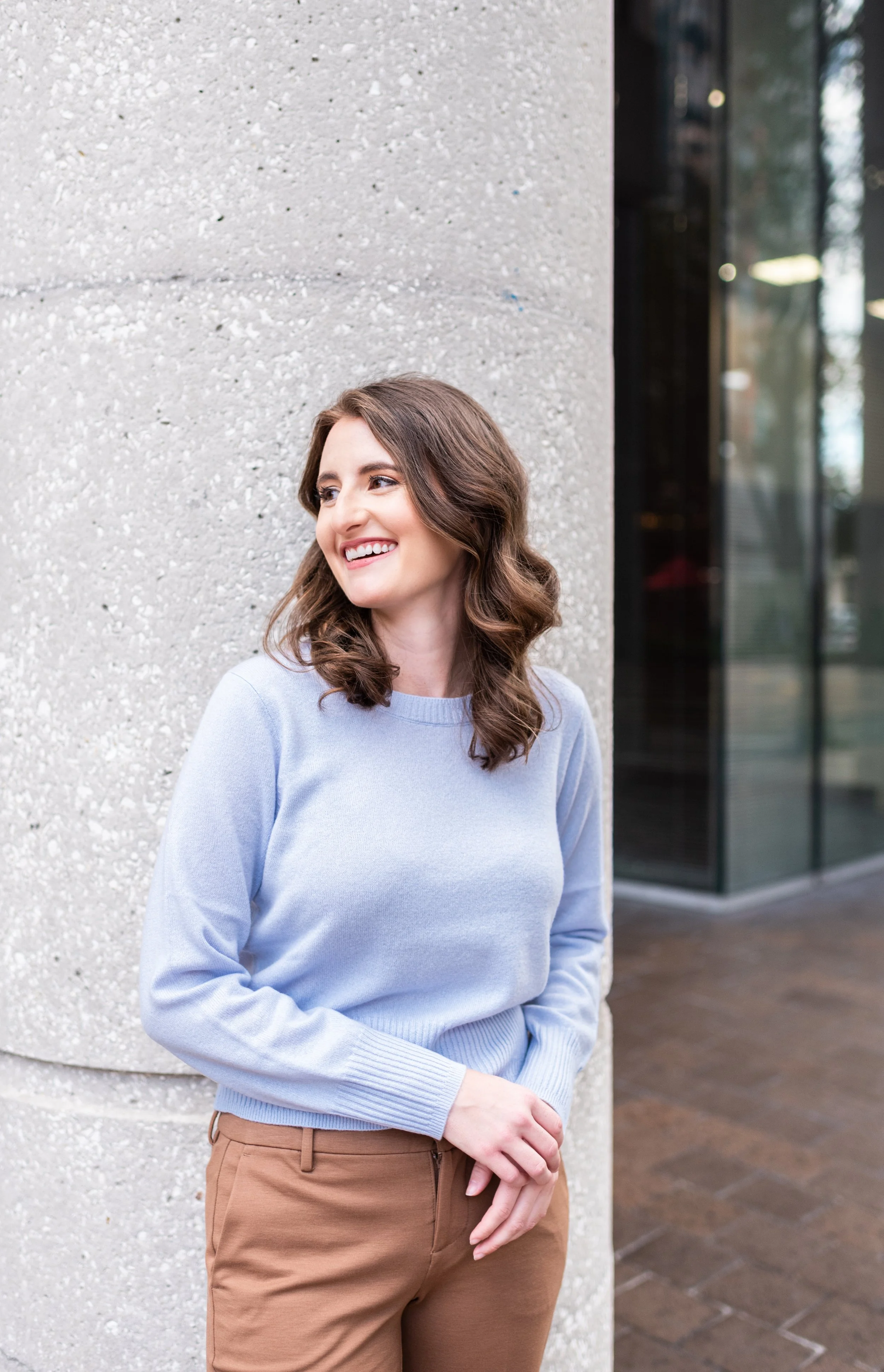 A woman with brown hair, wearing a light blue sweater and brown pants, leaning against a concrete wall outside a building, smiling.