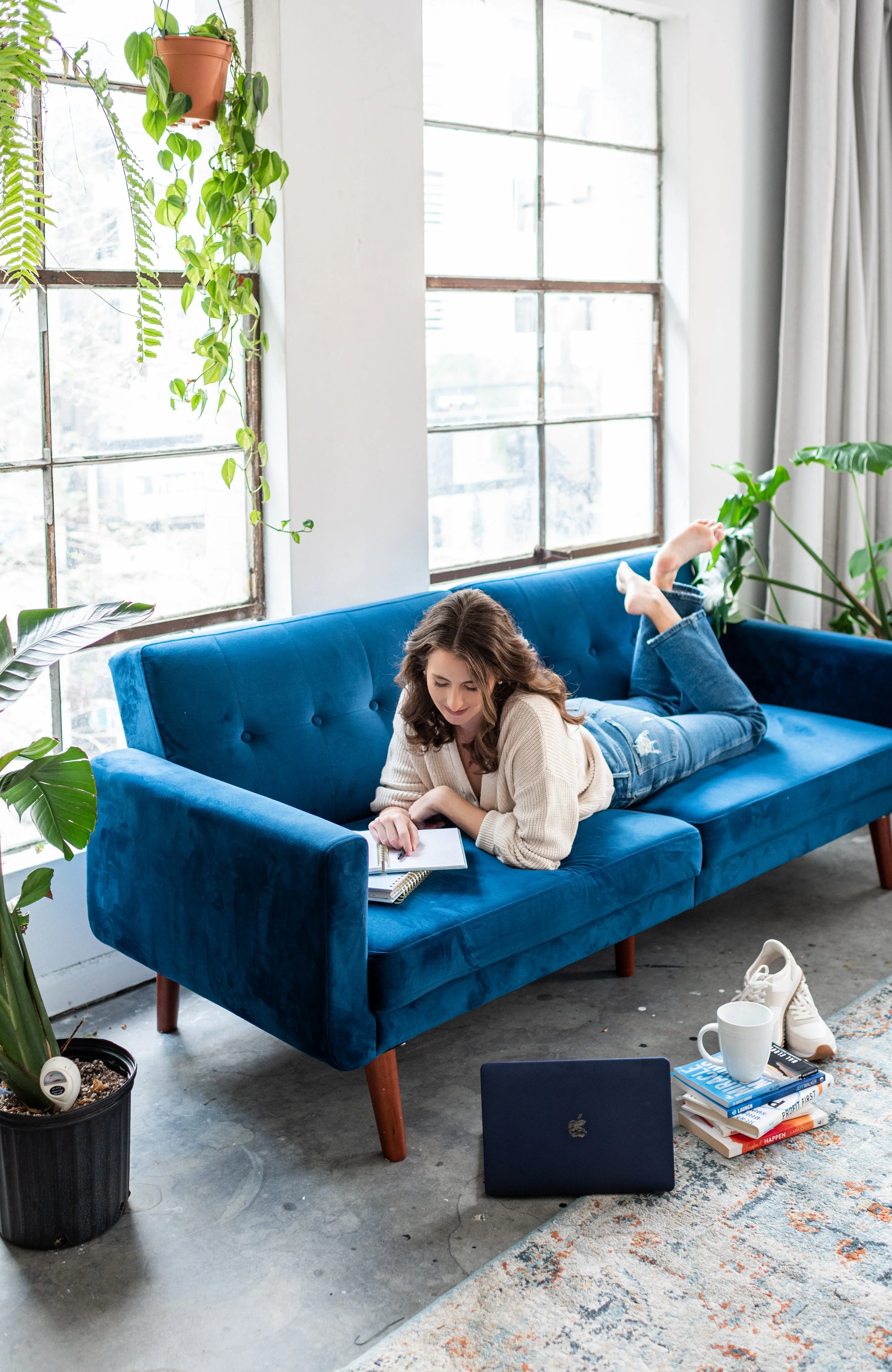 A woman lying on a blue velvet couch, writing in a notebook, with a laptop, a white mug, a pair of white sneakers, and a stack of books nearby in a bright room with large windows and green plants.