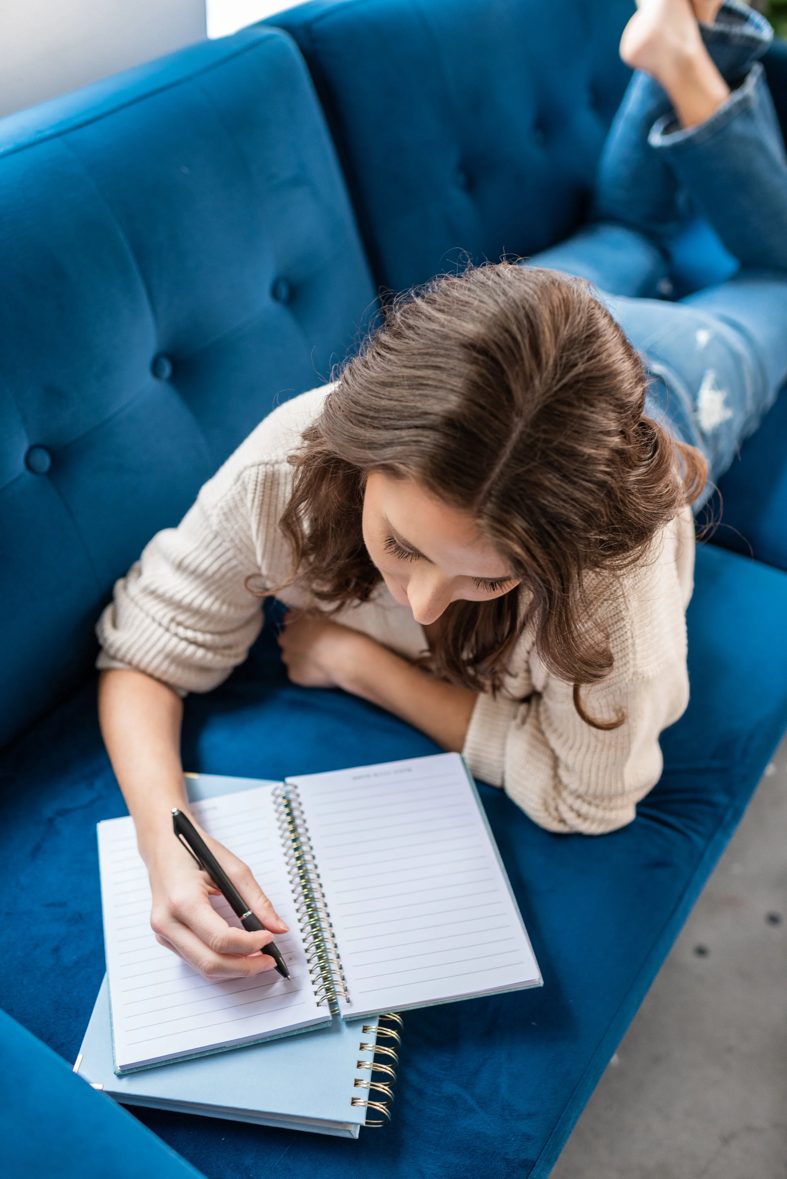 A woman lying on her stomach on a blue sofa, writing in a lined notebook with a black pen.