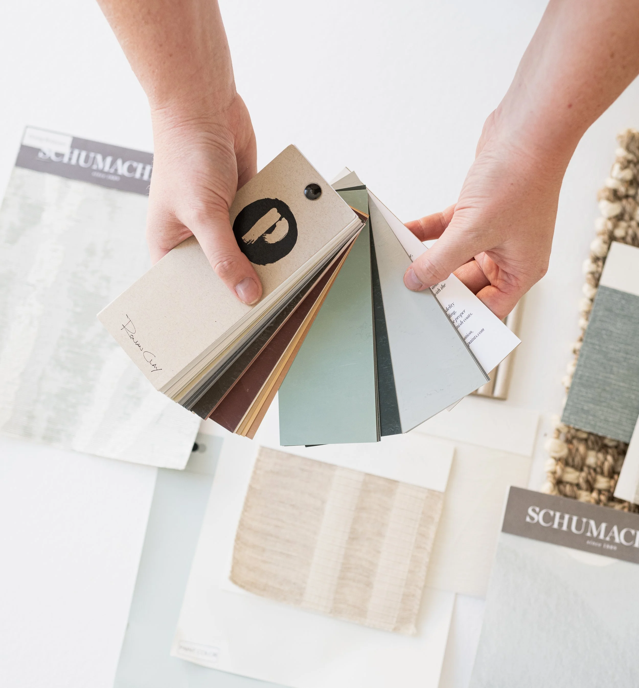 Person holding a fan deck of paint sample cards with various colors, with fabric swatches laid out on a white surface.