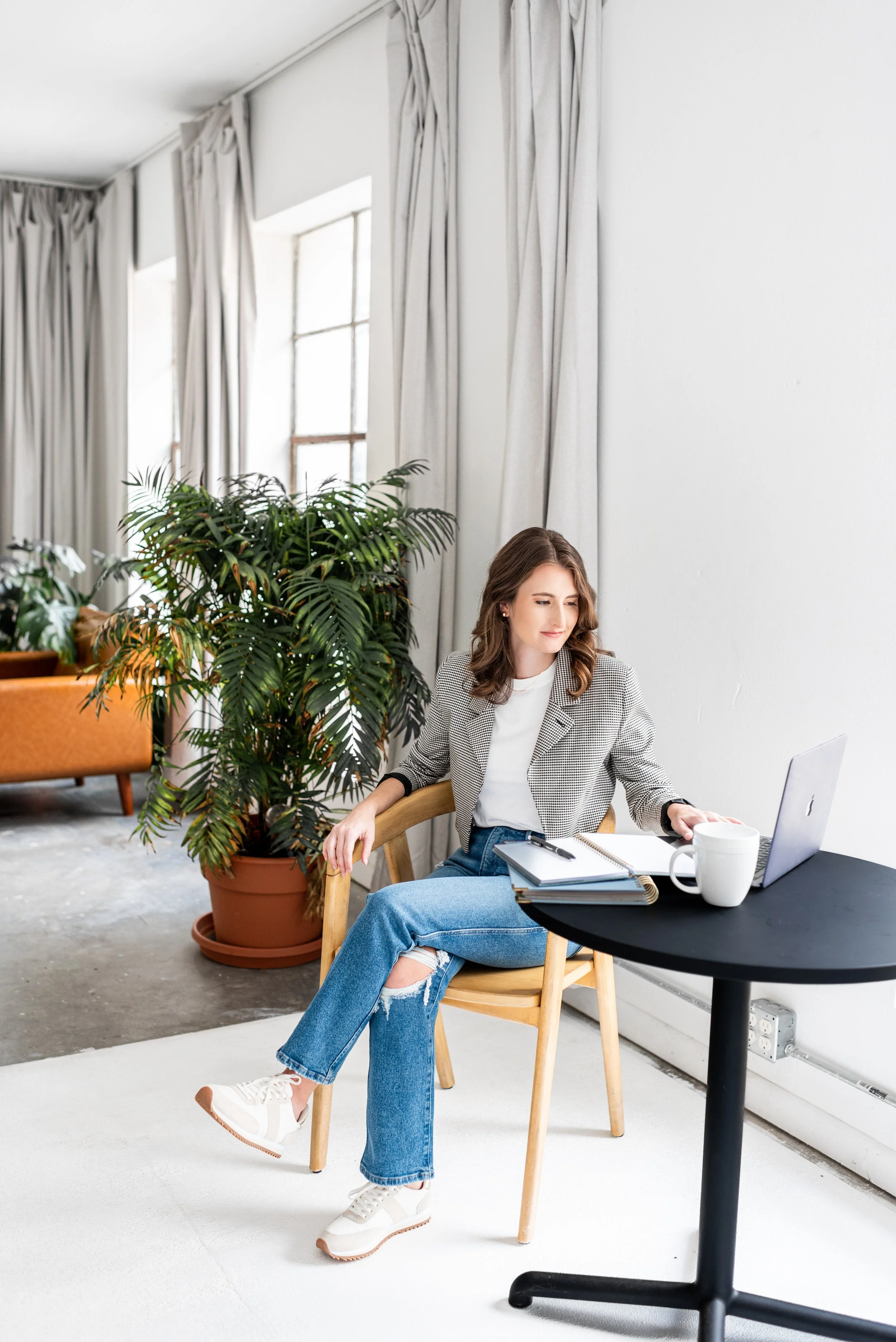A woman sitting at a black round table with a laptop, notebook, and coffee mug, in a bright room with large windows, white walls, white curtains, and a large potted plant.