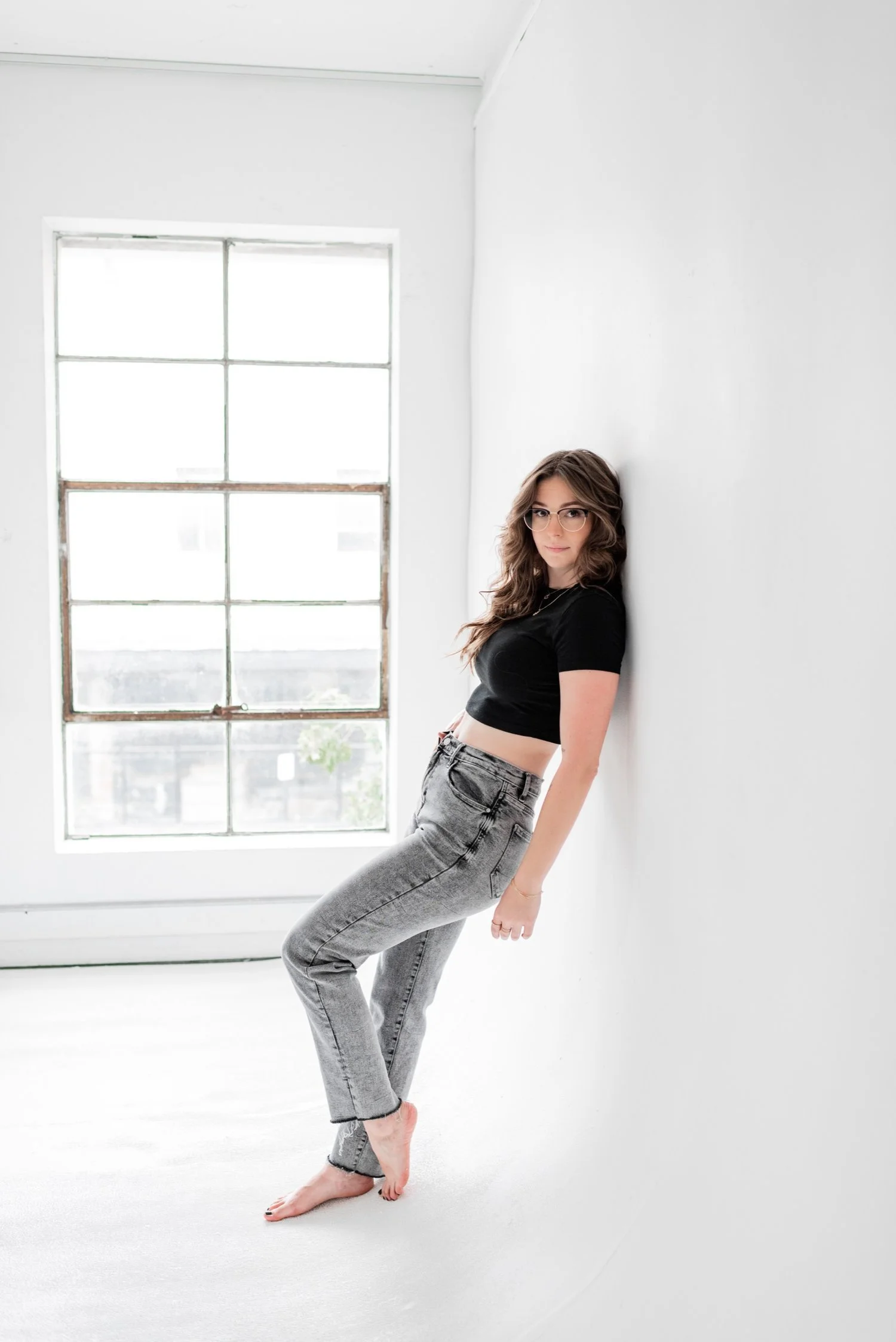 A young woman with glasses and wavy brown hair stands barefoot leaning against a white wall in front of a large window, wearing a black crop top and gray jeans in a bright, minimalist room.