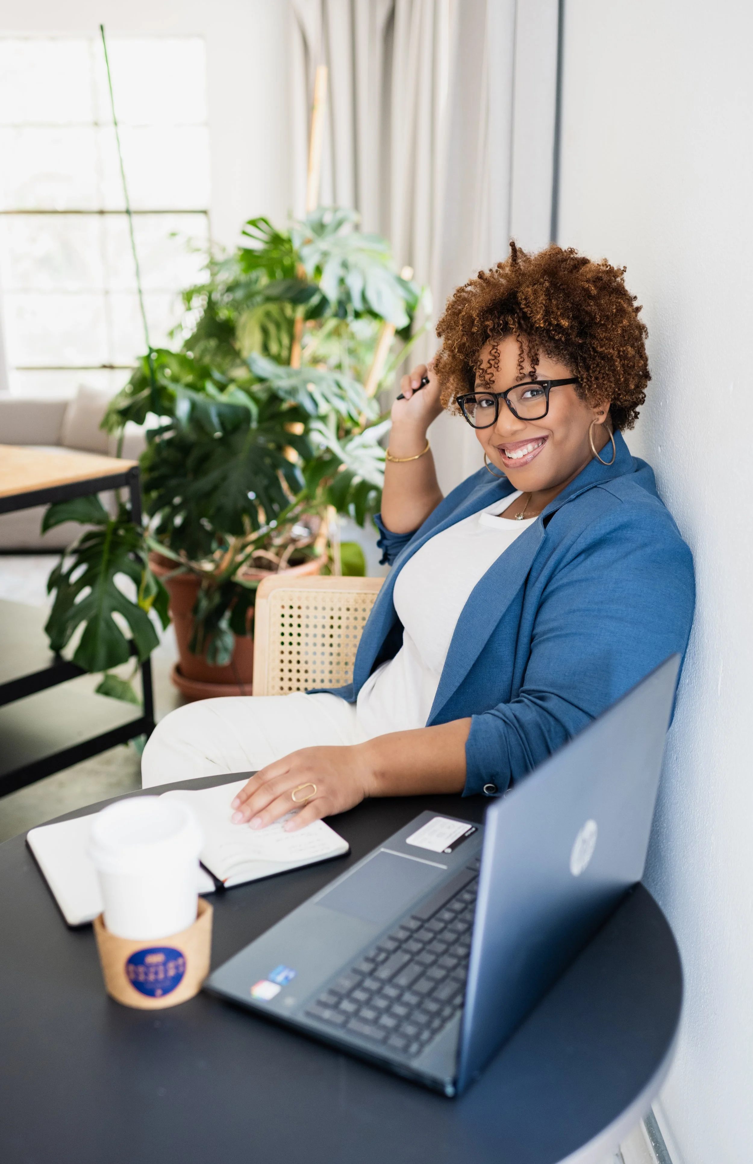 A woman with curly hair and glasses sitting at a table, smiling, with a laptop, notebook, and coffee in front of her, in a bright room with a large green plant in the background.