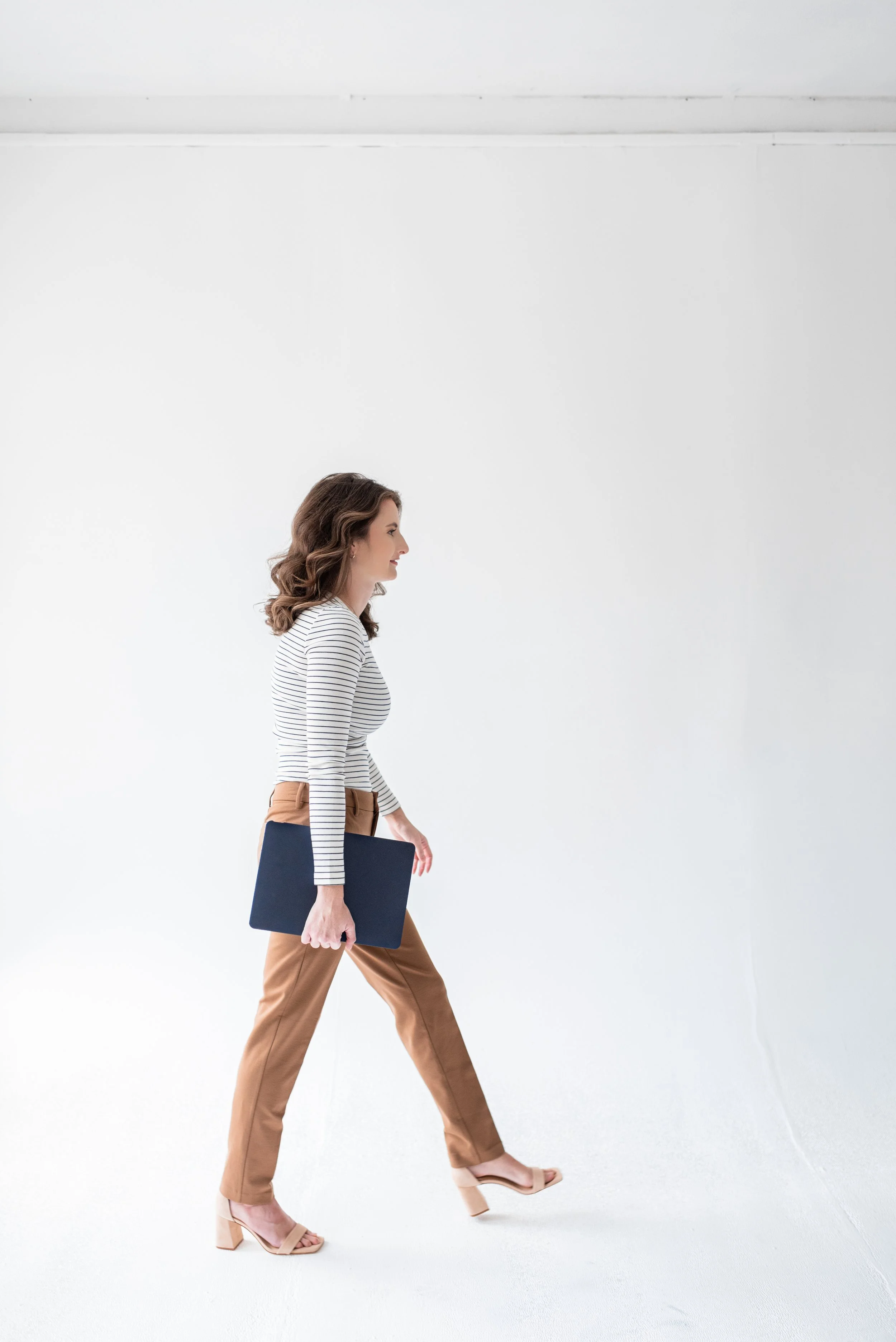 A woman walking in profile, holding a laptop, dressed in a striped top, tan pants, and nude heels, against a white background.