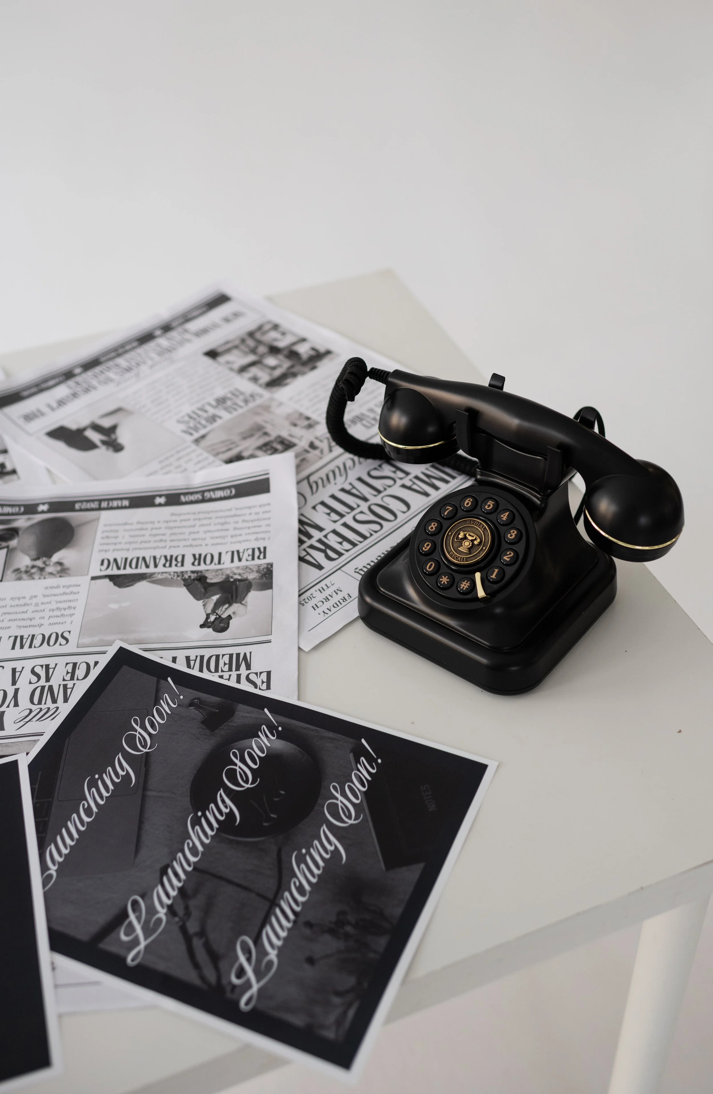 A black retro rotary phone on a white table surrounded by newspapers and a picture with the text 'Launching Soon!'.