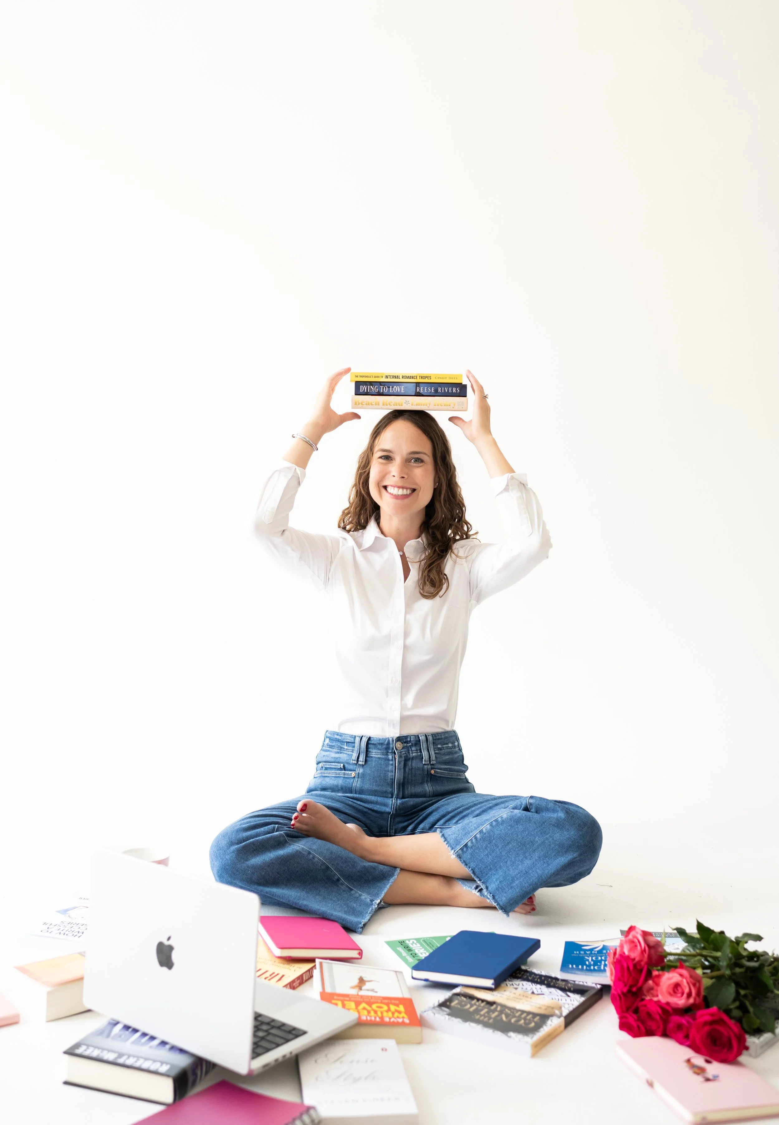 A woman sitting cross-legged on the floor holding three books above her head, smiling in front of a white background surrounded by books, a laptop, and a bouquet of pink roses.