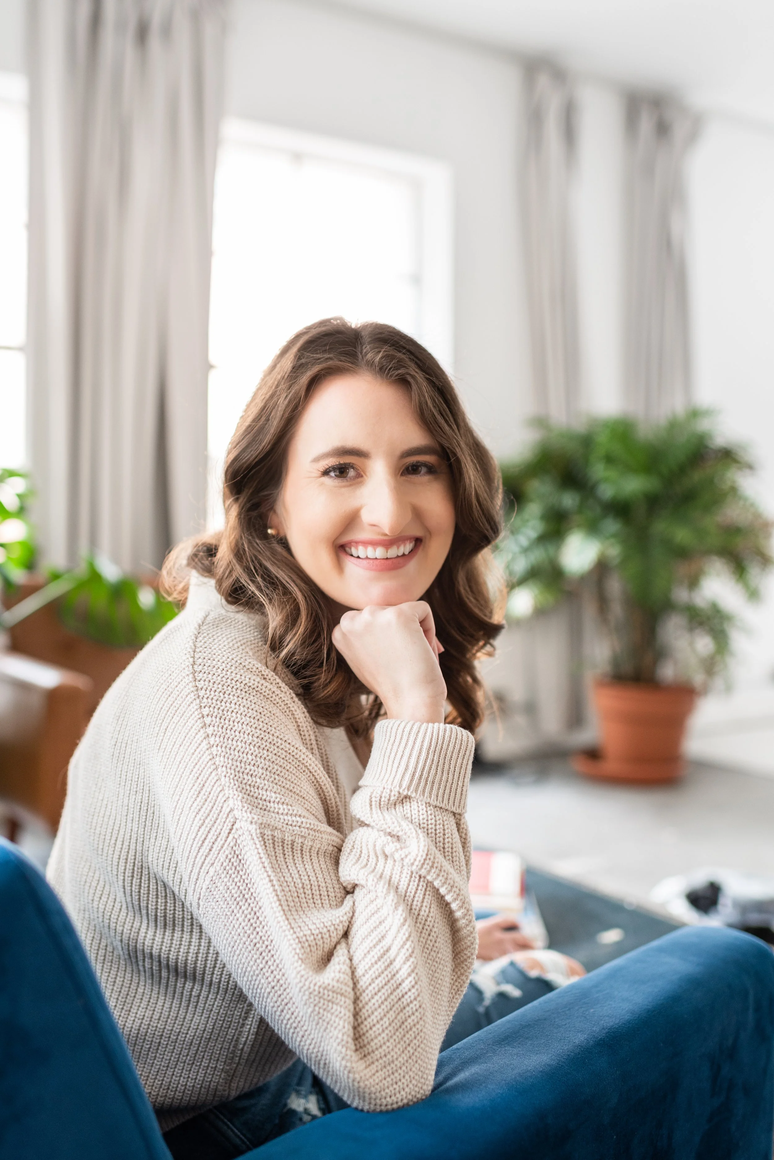 A smiling woman sitting on a blue couch in a bright living room, with a large potted plant and window with curtains in the background.