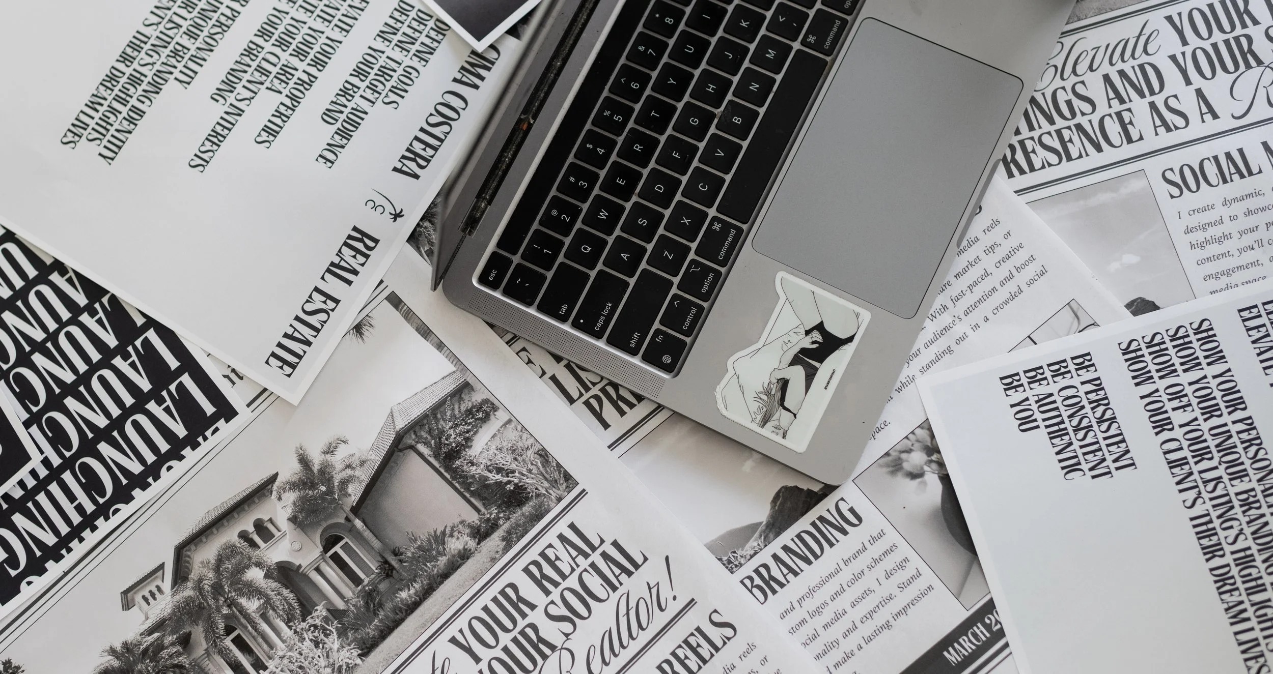 A flat lay of a silver laptop with black keyboard, surrounded by printed marketing materials and brochures featuring black and white text and images, with a small illustration sticker near the laptop.