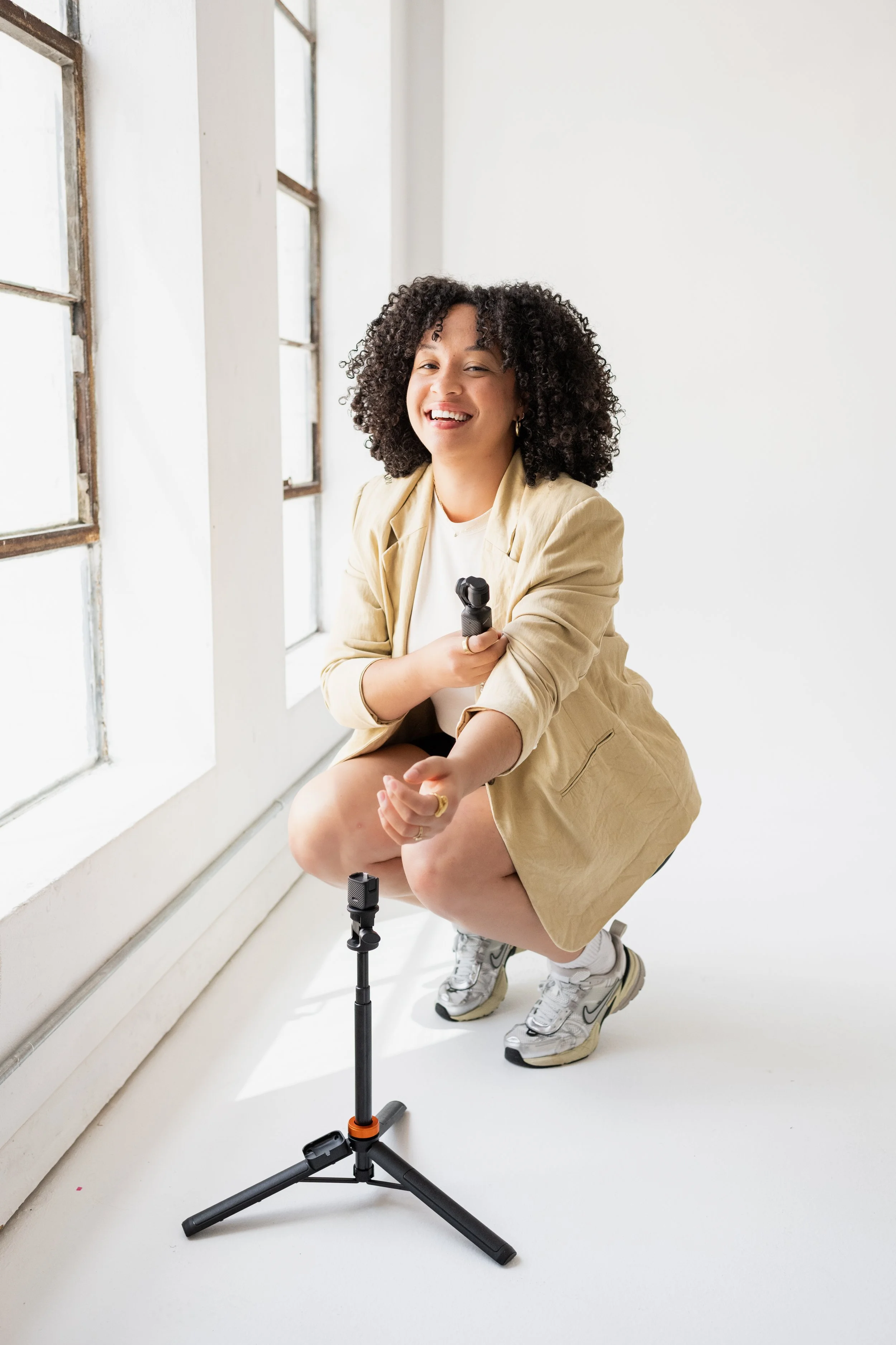 A woman crouching near a window with a smile, holding a microphone, and a small tripod stand in front of her, in a bright white space.
