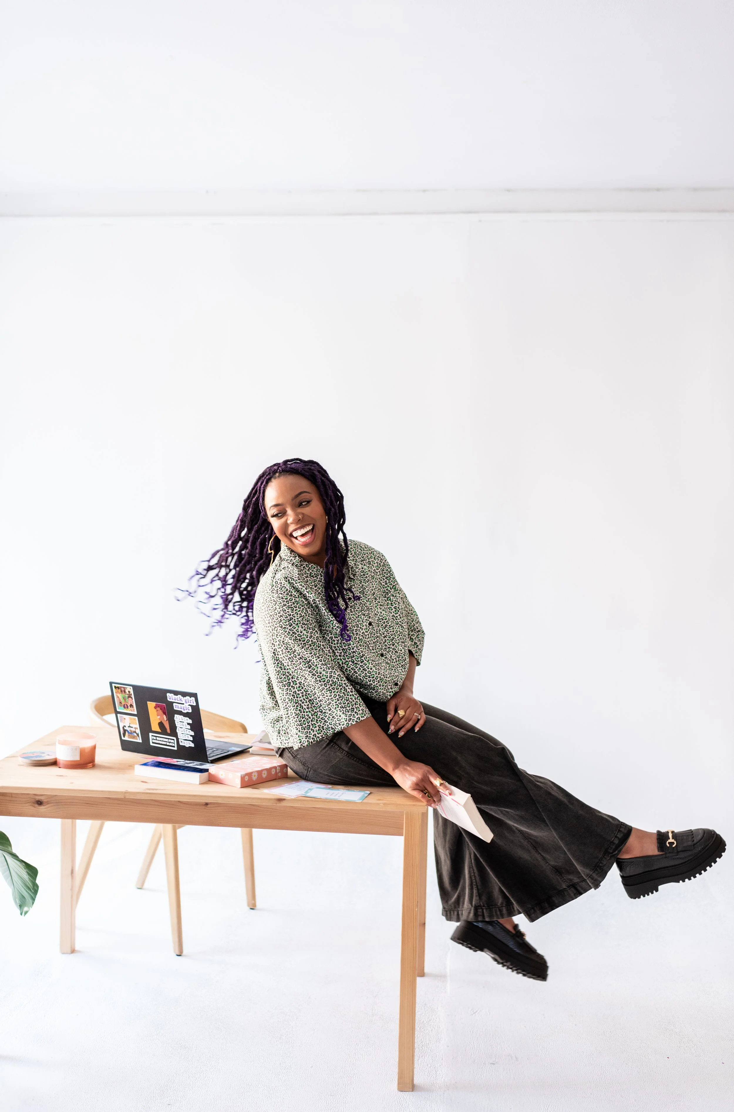 A woman with purple dreadlocks sitting sideways on a wooden table, smiling, holding papers with her feet off the ground, with a white background and a laptop and books on the table.