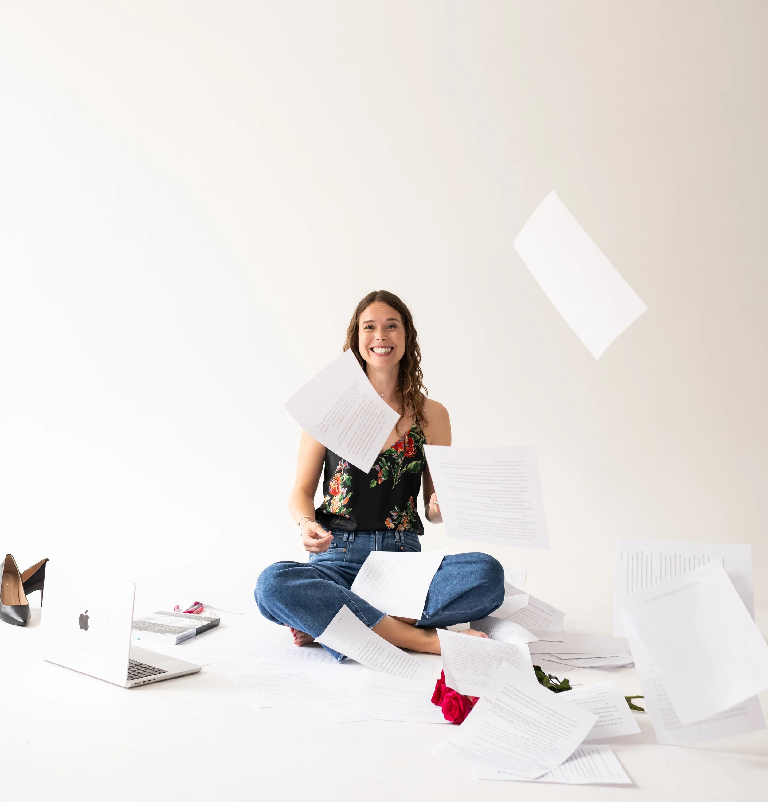 A woman sitting cross-legged on the floor, smiling, with papers flying around her, a laptop, shoes, a notebook, and roses nearby.