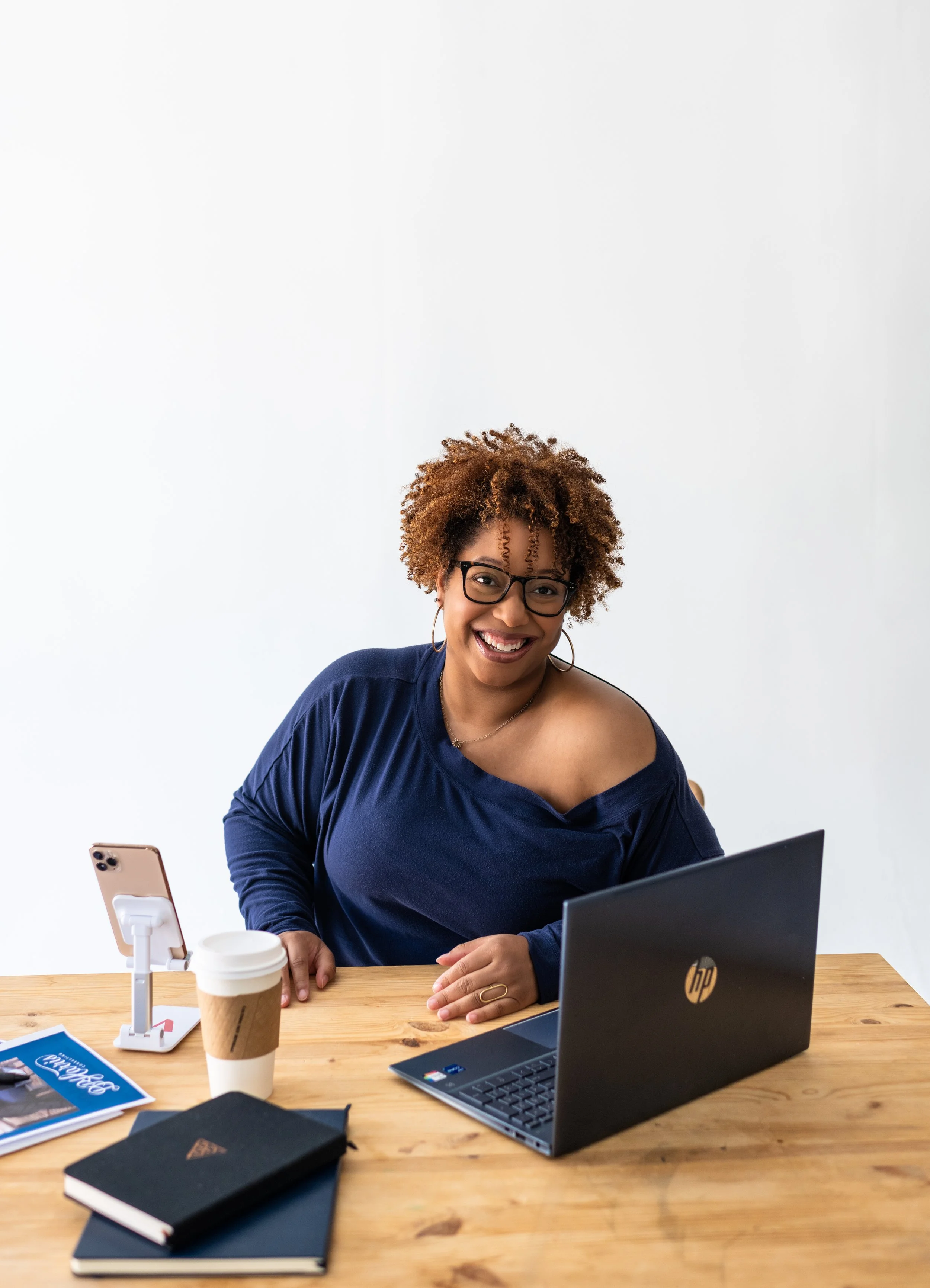 A woman with curly hair and glasses smiling at a wooden desk with a laptop, coffee cup, smartphone on a stand, and notebooks.