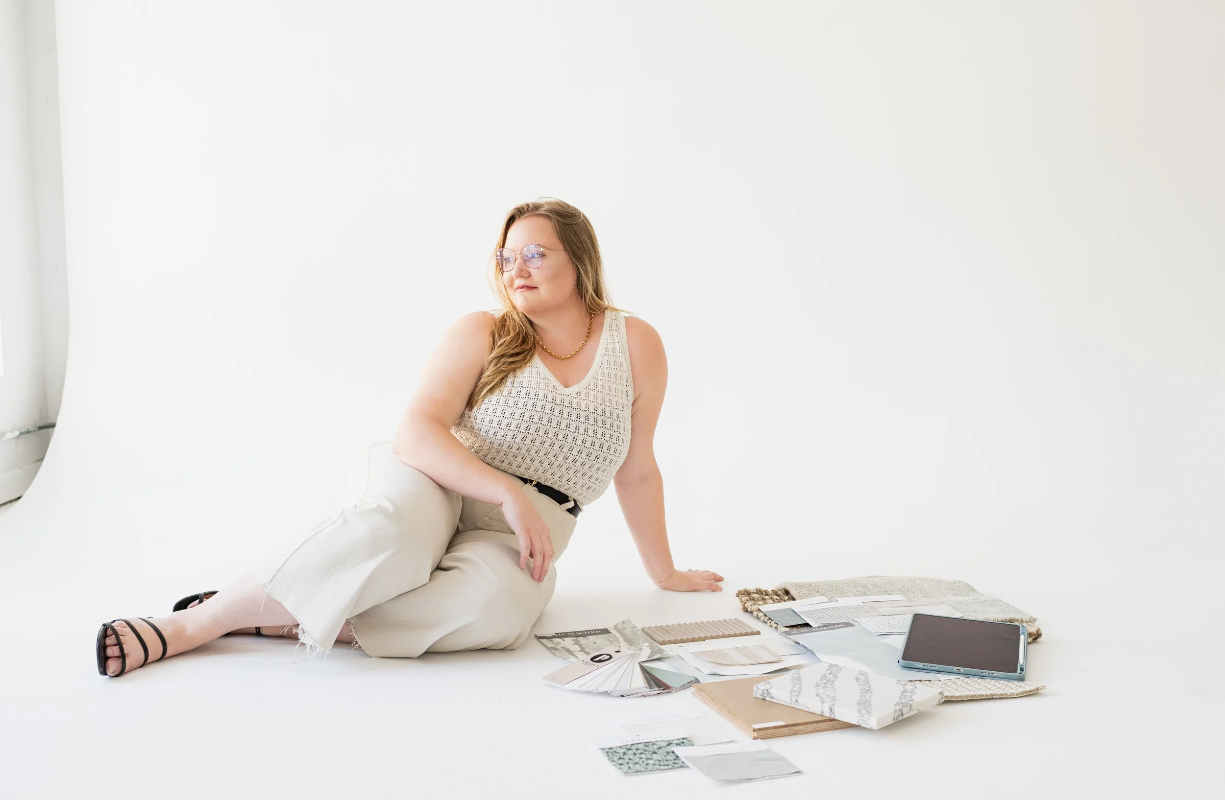 A woman with long hair, glasses, and a sleeveless top sitting on the floor surrounded by fabric samples, color swatches, and a tablet in a bright, white studio.