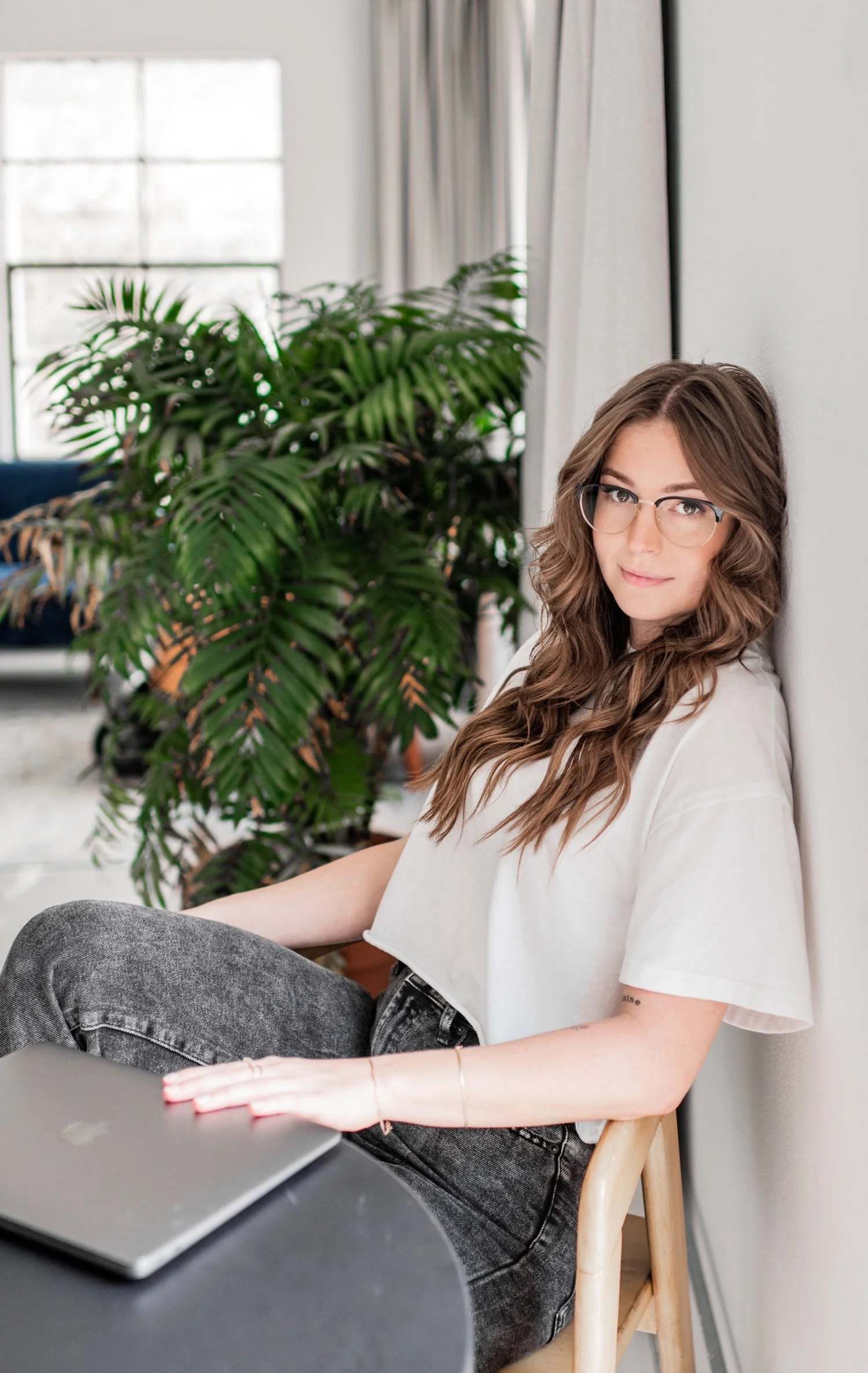 A young woman with wavy brown hair and glasses sitting on a wooden chair next to a white wall, with a laptop in front of her and a large green leafy plant behind her in a bright indoor space.