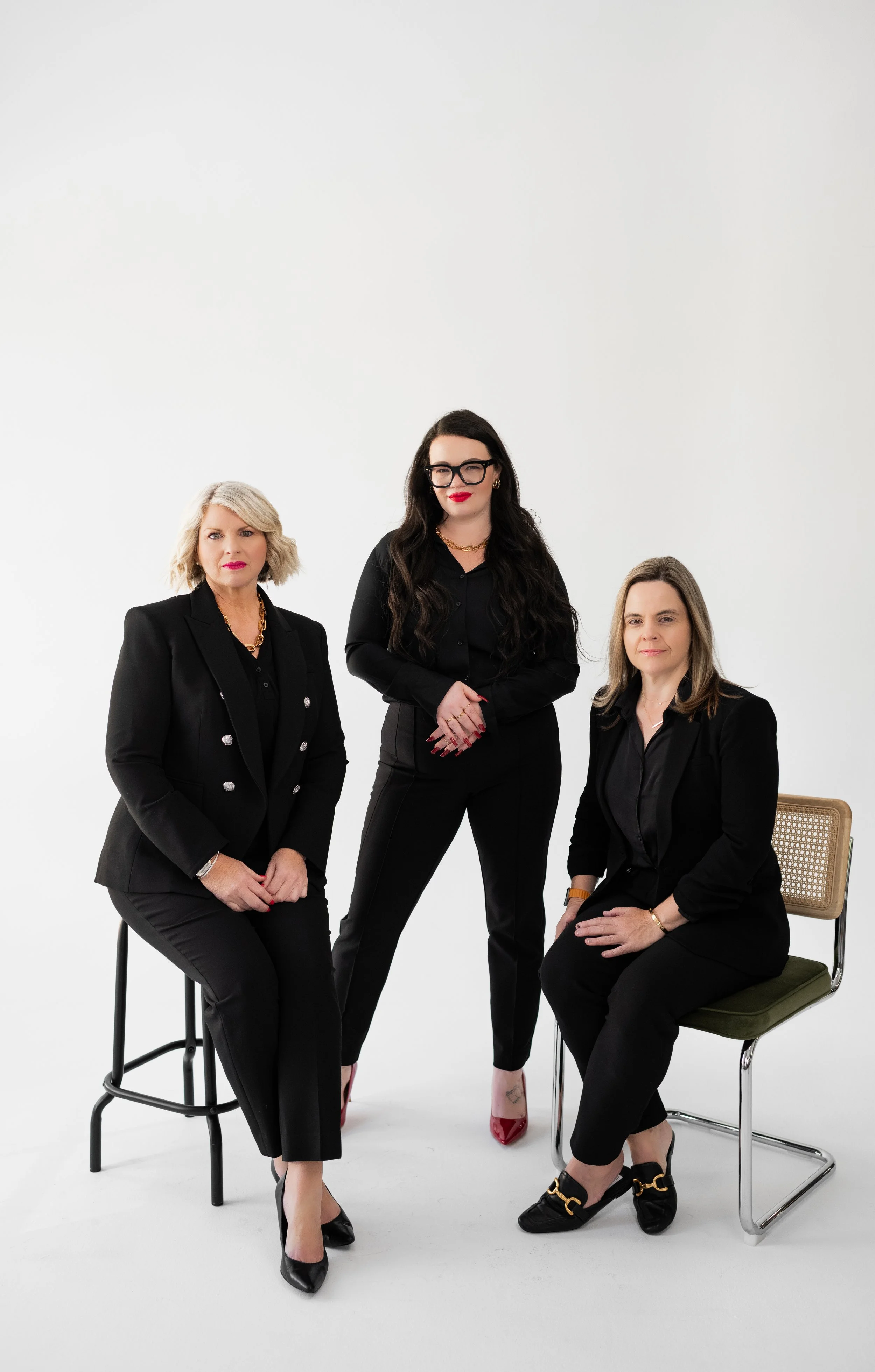 Three women dressed in black suits posing in a studio with a white background, two sitting on chairs and one standing.