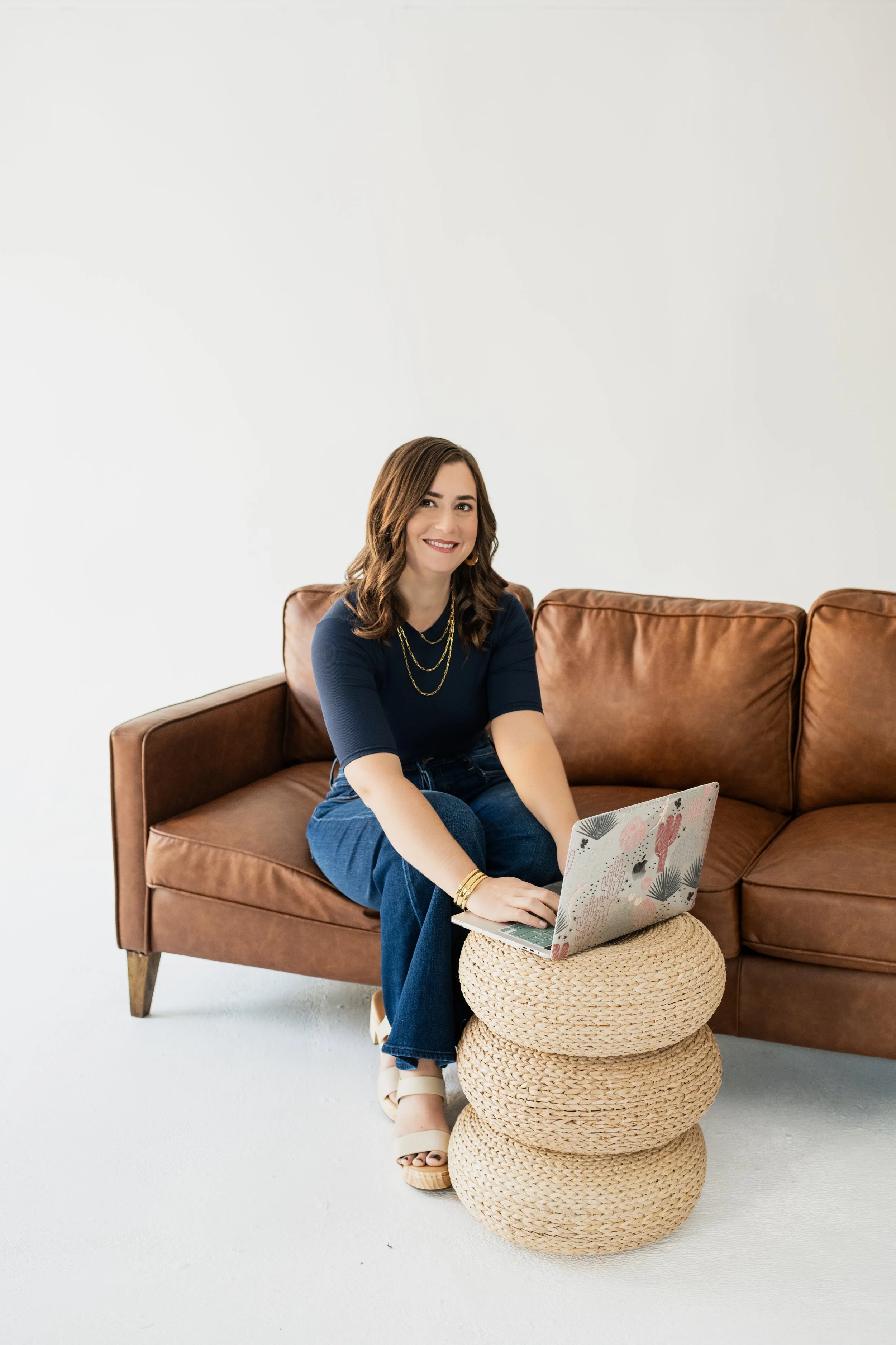Woman sitting on a brown leather sofa, smiling, working on a laptop placed on a stack of three round woven stools, in a bright room with white walls.