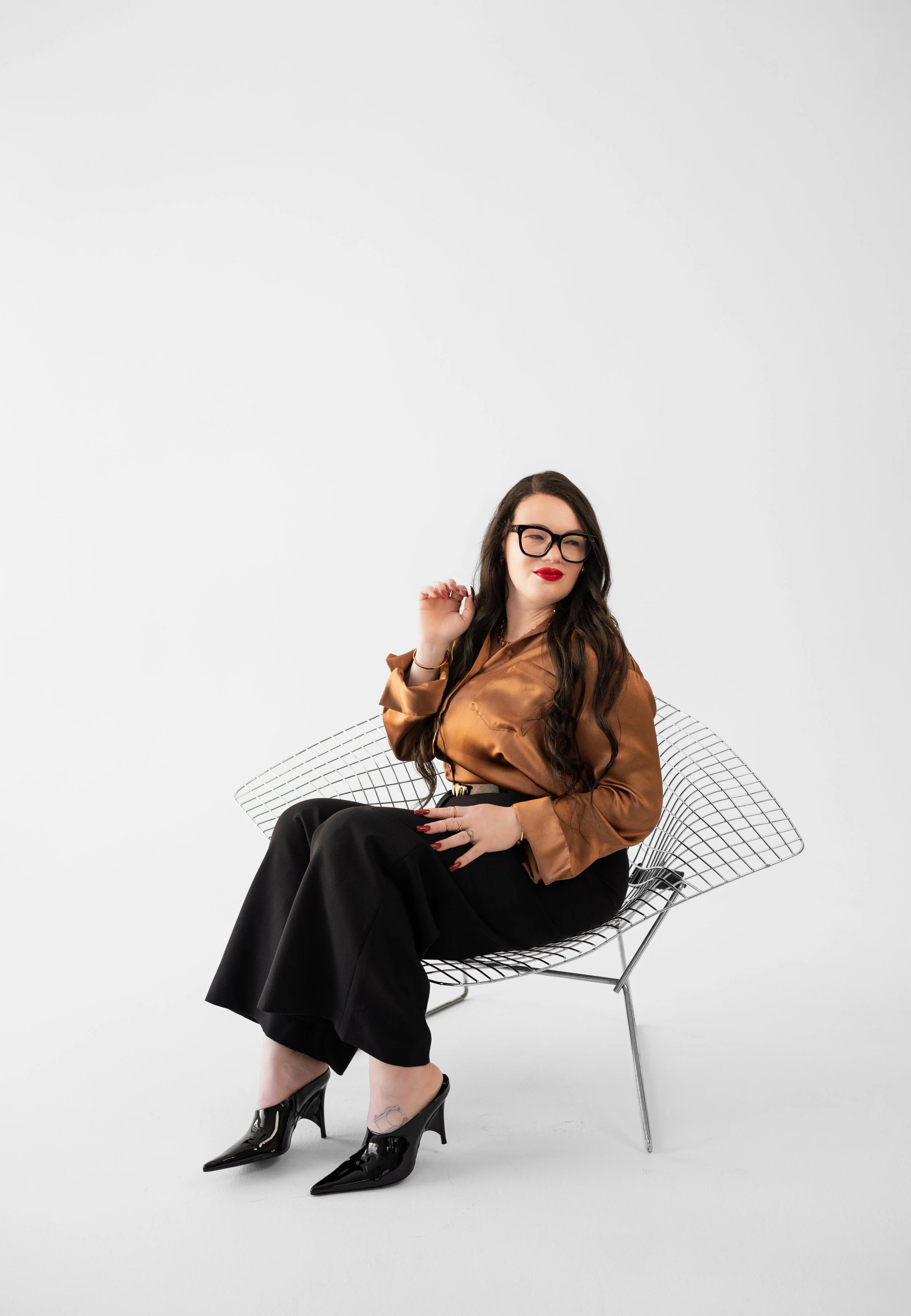 A woman with long dark hair, glasses, red lipstick, wearing a brown satin blouse, black wide-leg pants, and black high heels, sitting on a wire chair against a plain white background.