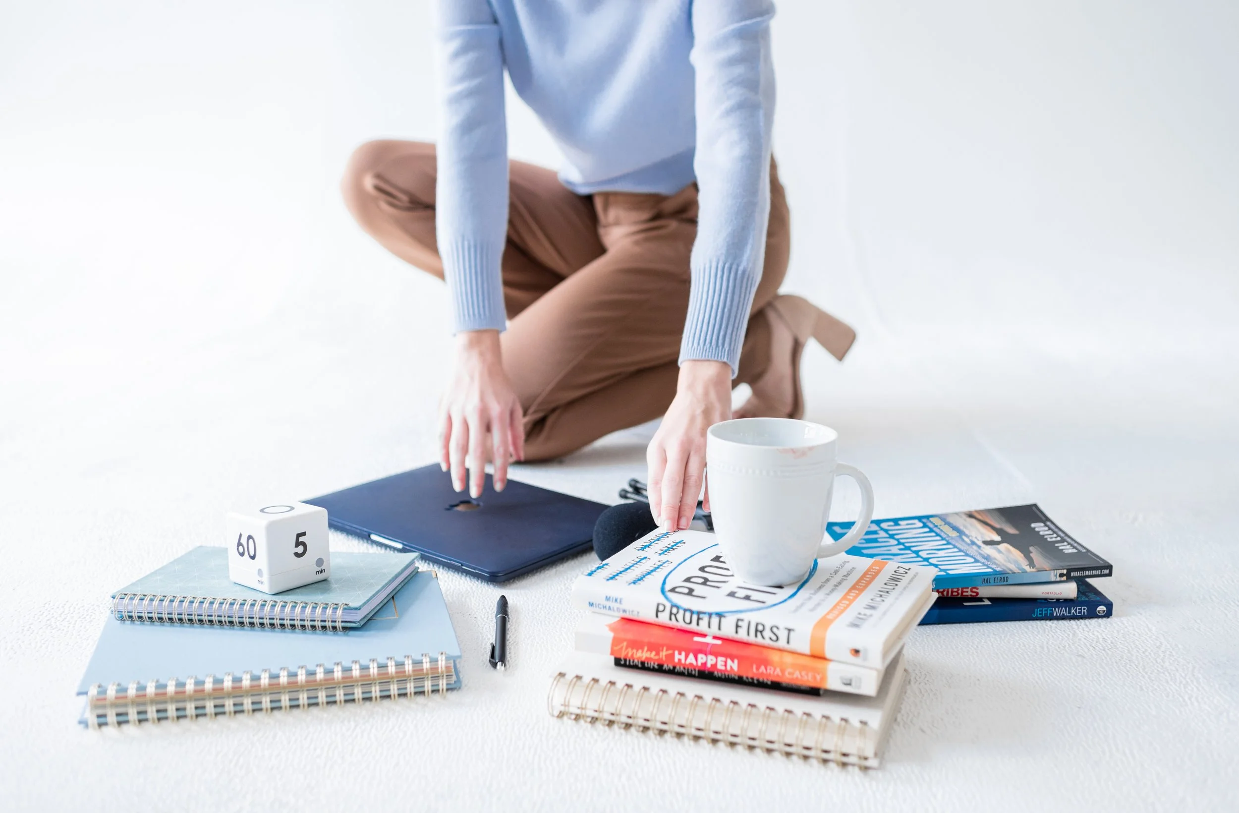Person in a blue sweater and brown pants kneeling on a white surface, surrounded by notebooks, books, a coffee mug, and a small white dice.