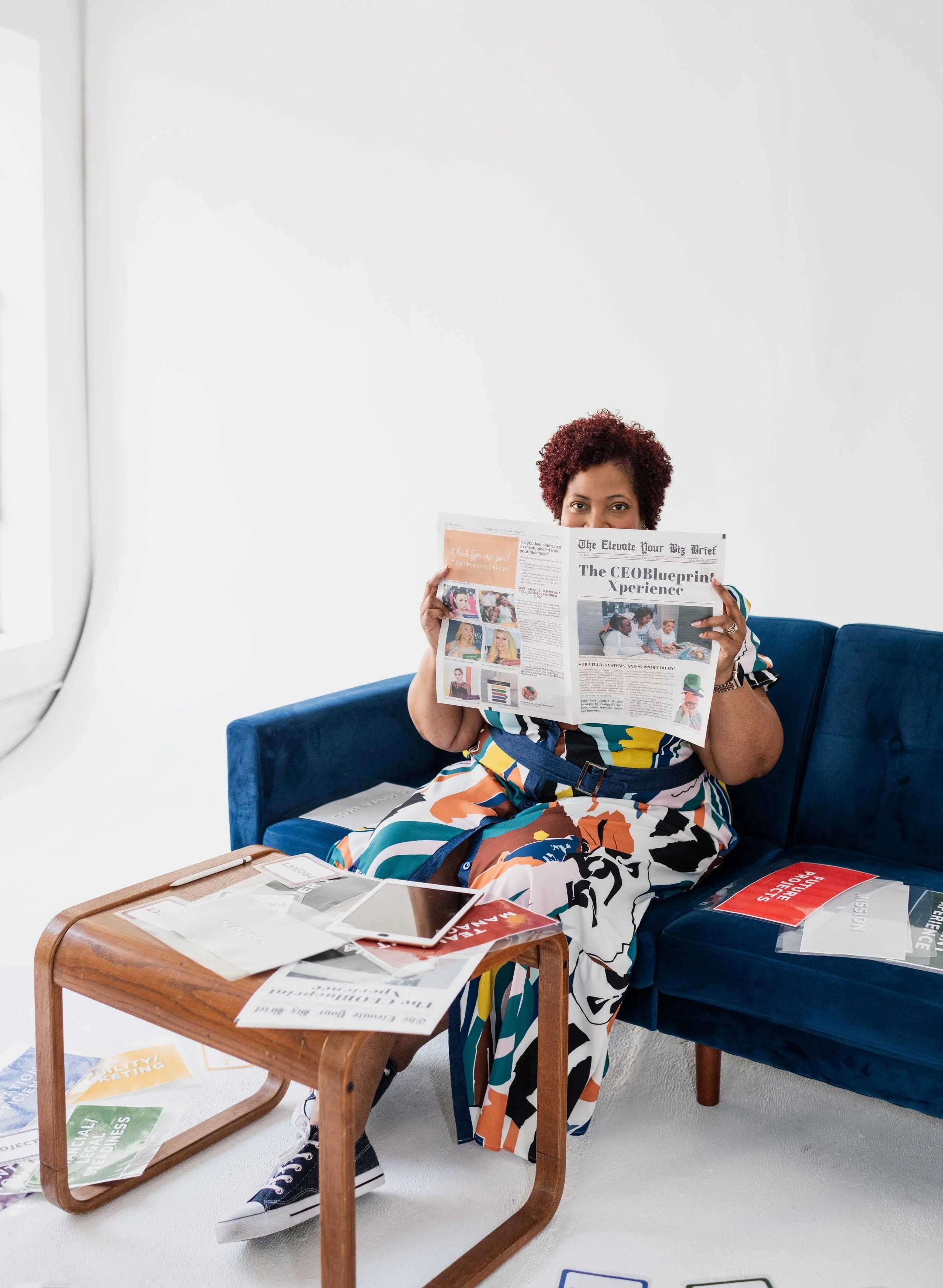 A woman with curly hair sitting on a blue sofa, reading a newspaper in a bright room with a white wall. There are magazines, a tablet, and papers on a wooden coffee table in front of her.