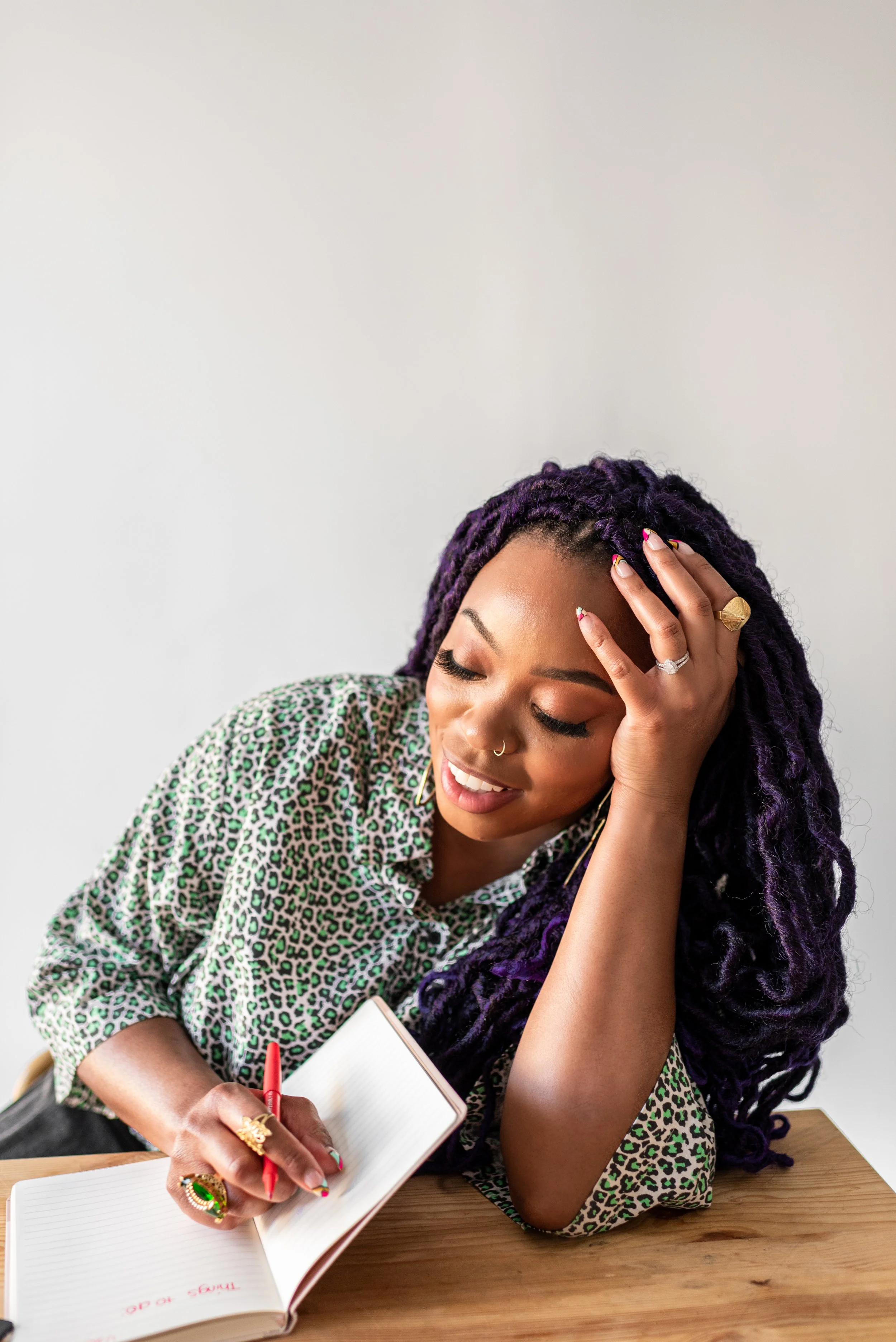 A woman with purple dreadlocks, wearing a leopard print shirt, is smiling while writing in a notebook on a wooden table.