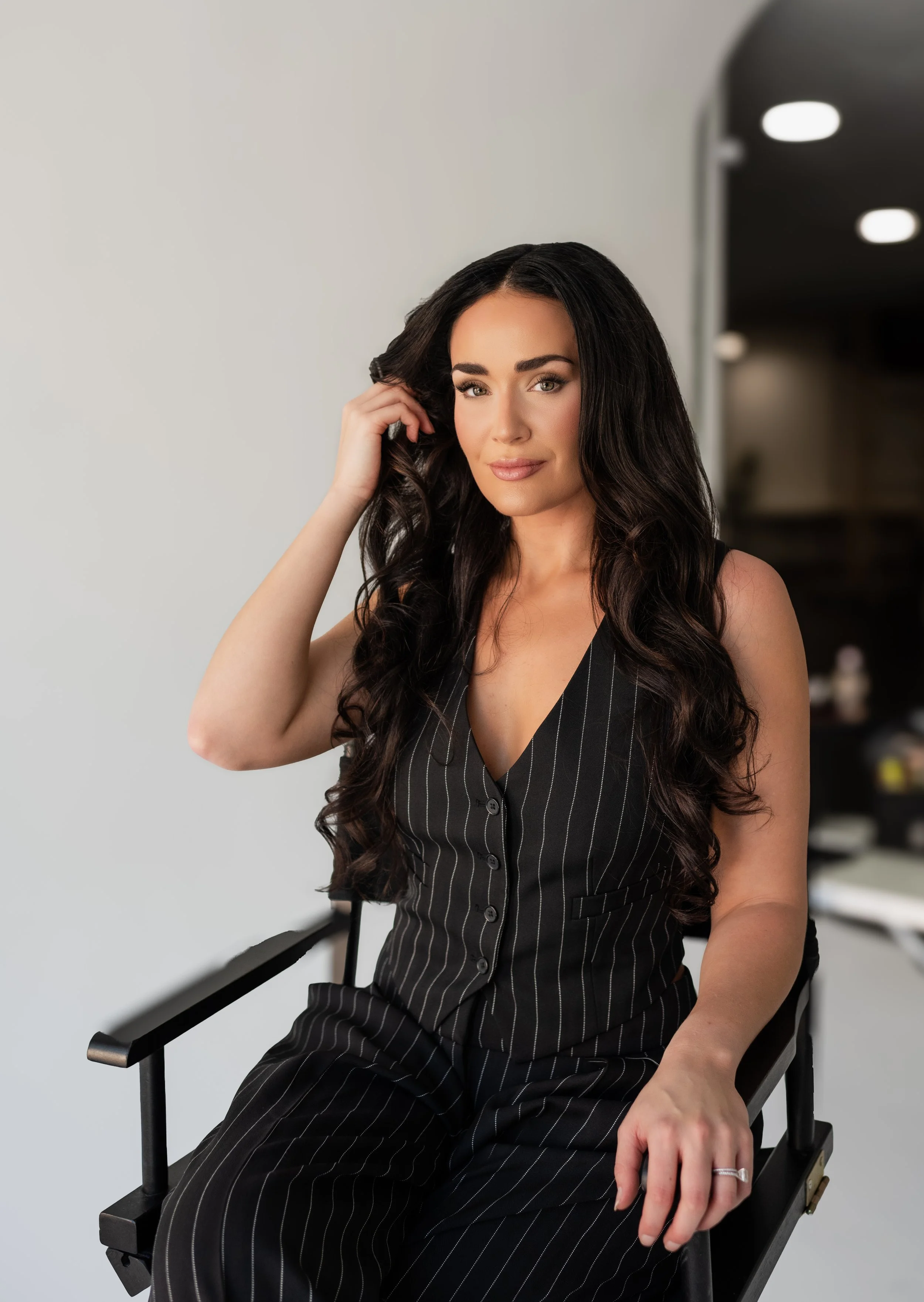 Portrait of a woman with long dark wavy hair, sitting on a black director's chair in a modern indoor setting, wearing a black pinstripe sleeveless suit, smiling softly.