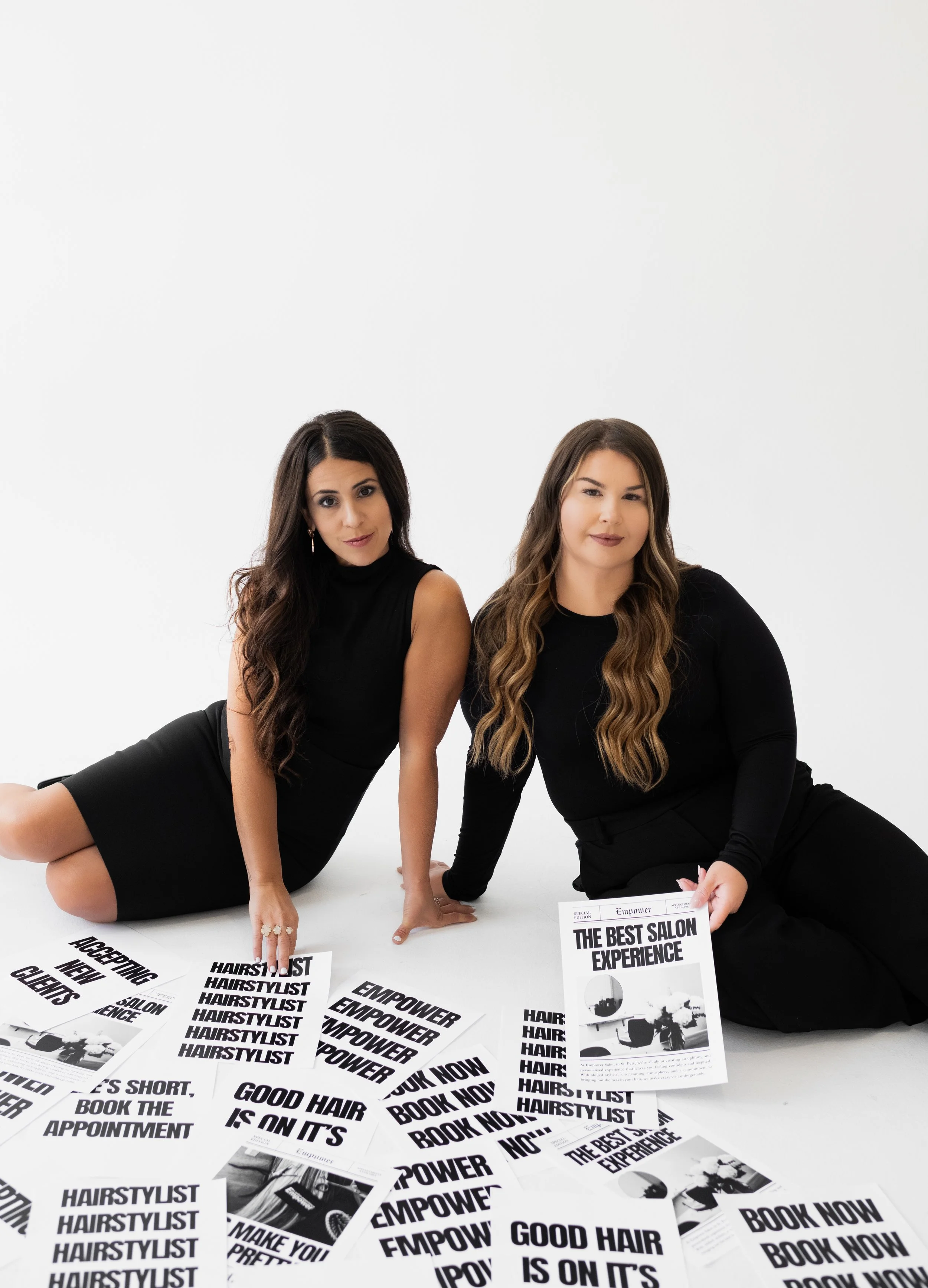 Two women sitting on the floor surrounded by black-and-white printed flyers promoting a hair salon experience and appointment booking.