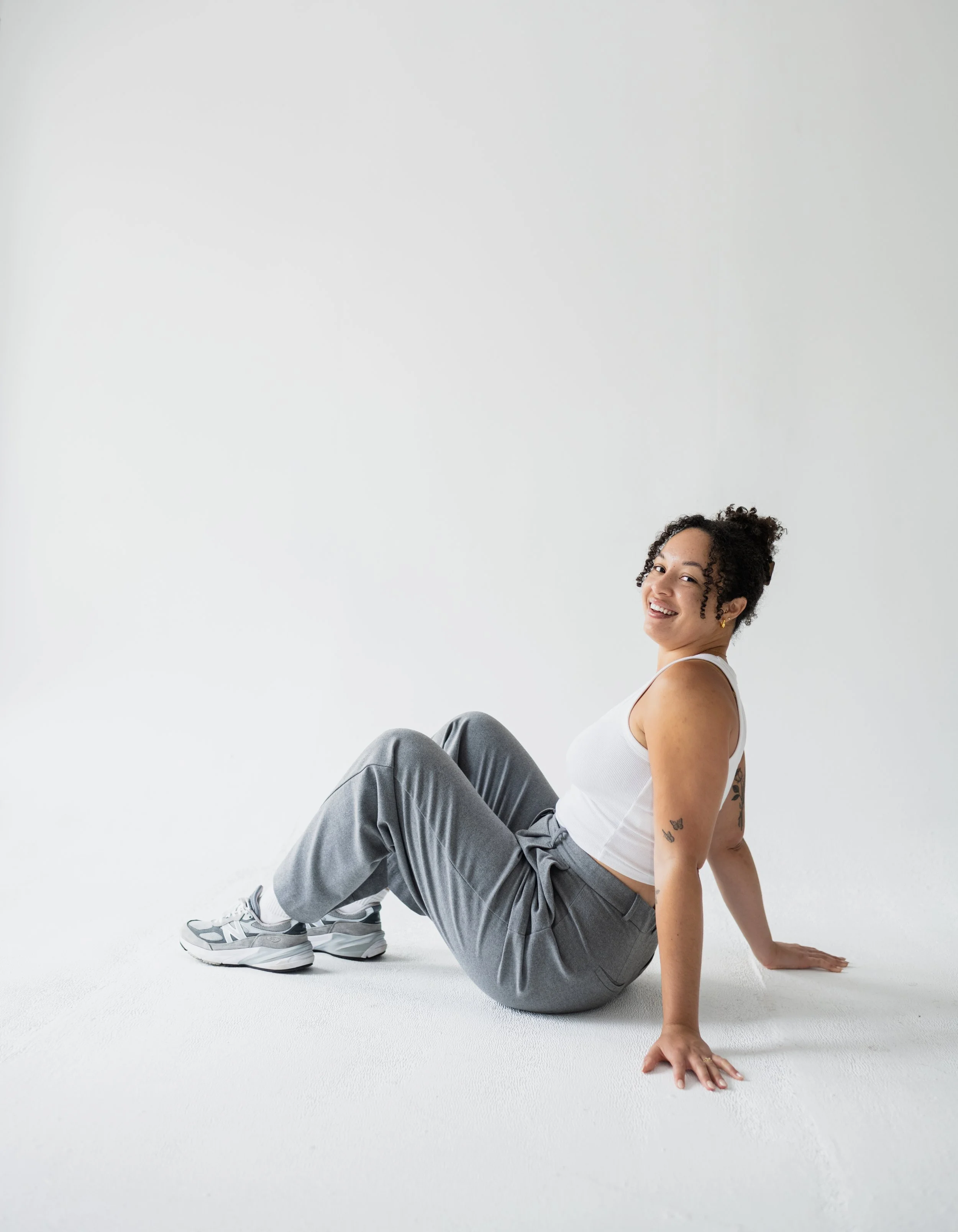 A woman wearing a white tank top, gray sweatpants, and sneakers, smiling while sitting on a white floor against a plain white background.