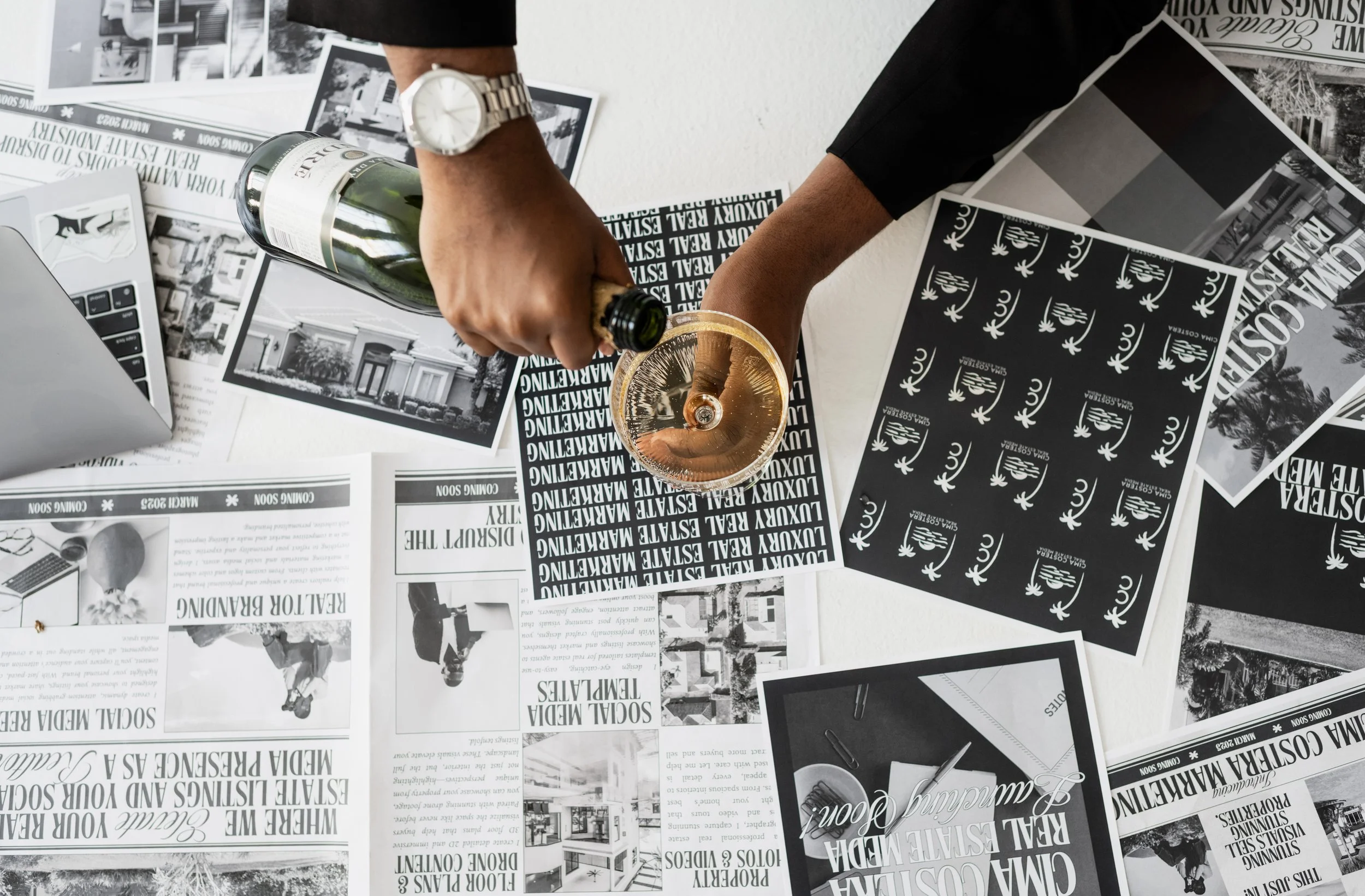 A person pouring champagne into a glass on a table covered with black-and-white printed papers and photographs, with an open laptop and a bottle of champagne.