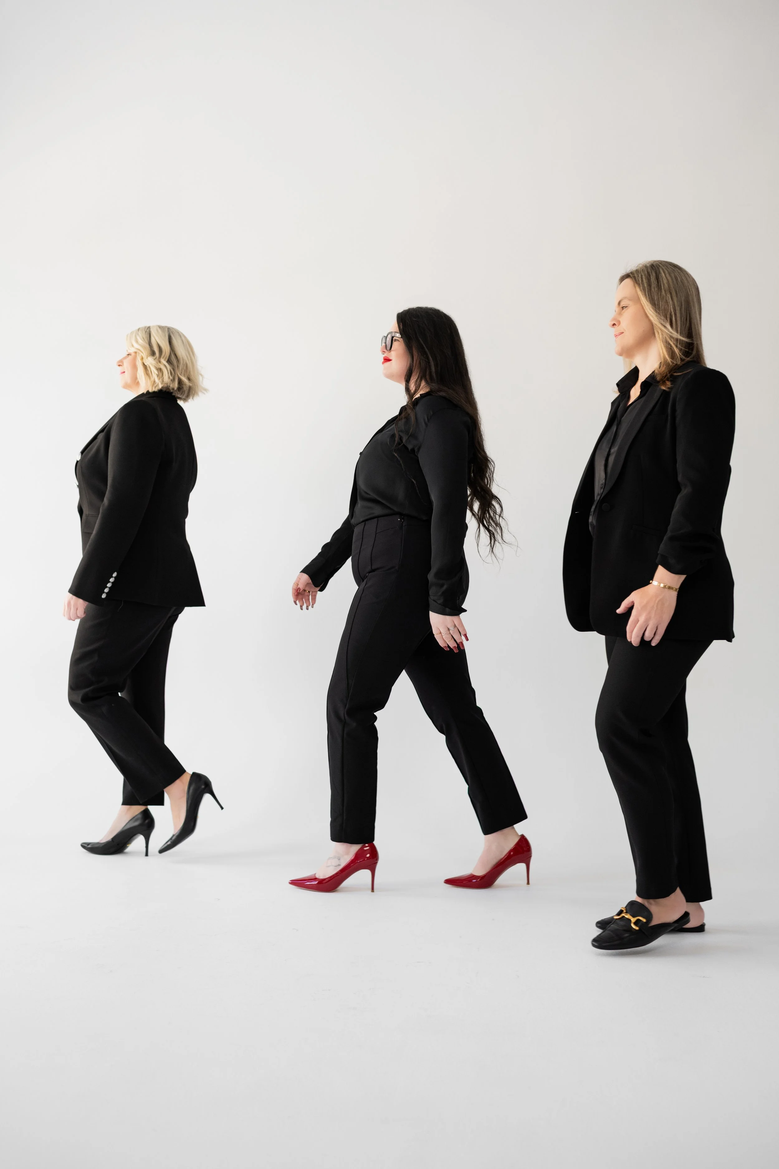 Three women in black business suits walking in a white studio, wearing high heels.