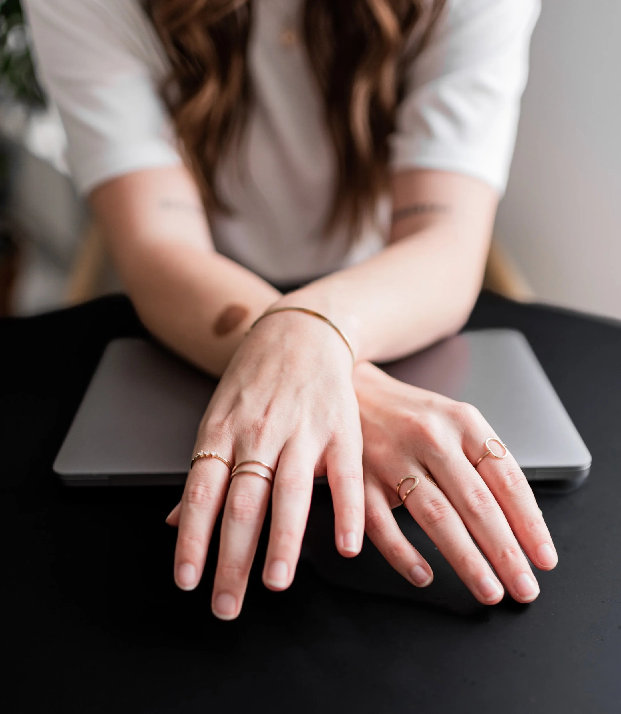 A woman with long brown hair wearing a white top, showing her adorned hands with several rings, resting on a closed laptop.