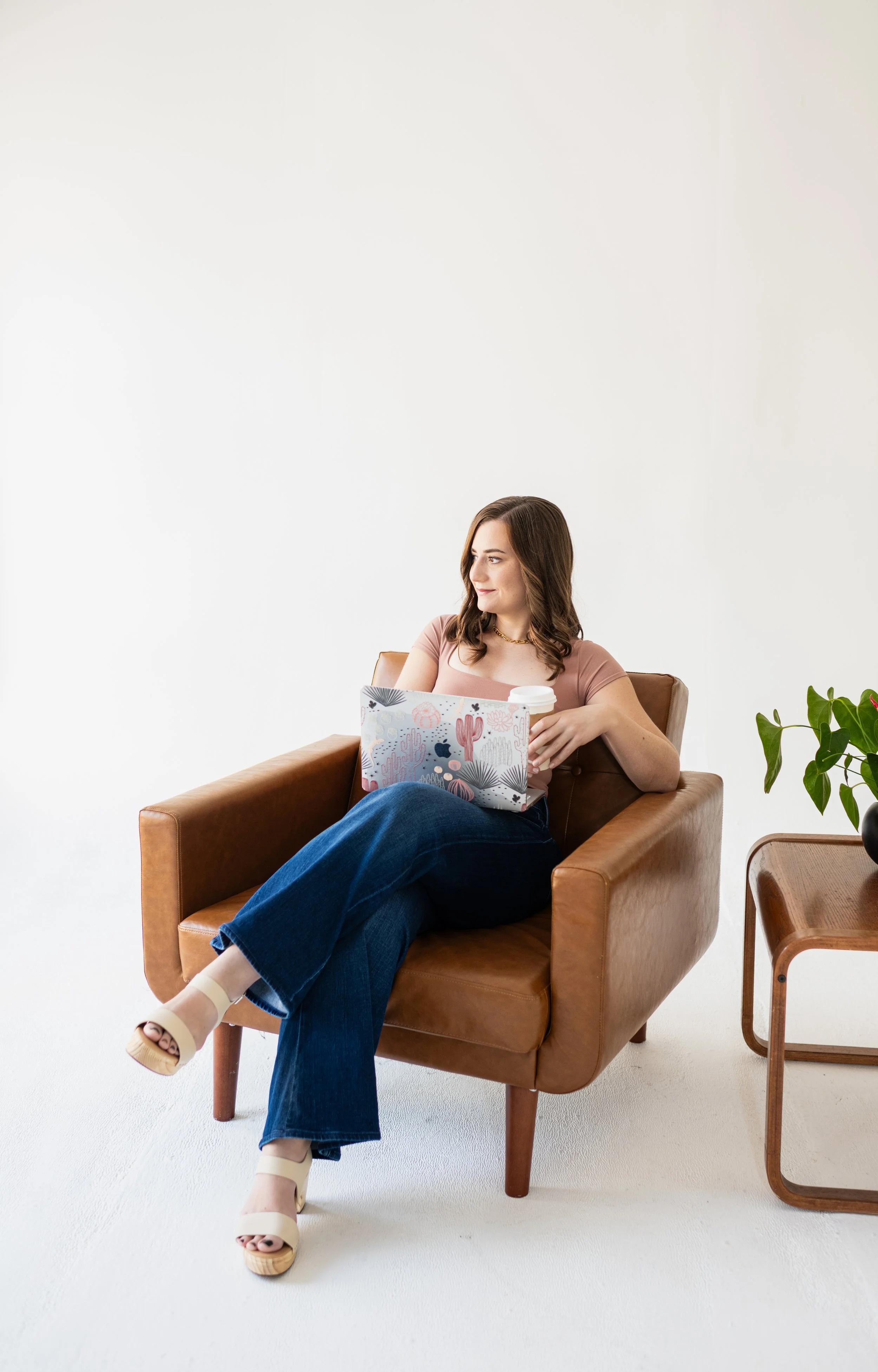 Woman sitting on a leather chair with a laptop and coffee, next to a small table with a plant, in a minimalist room with a white wall.