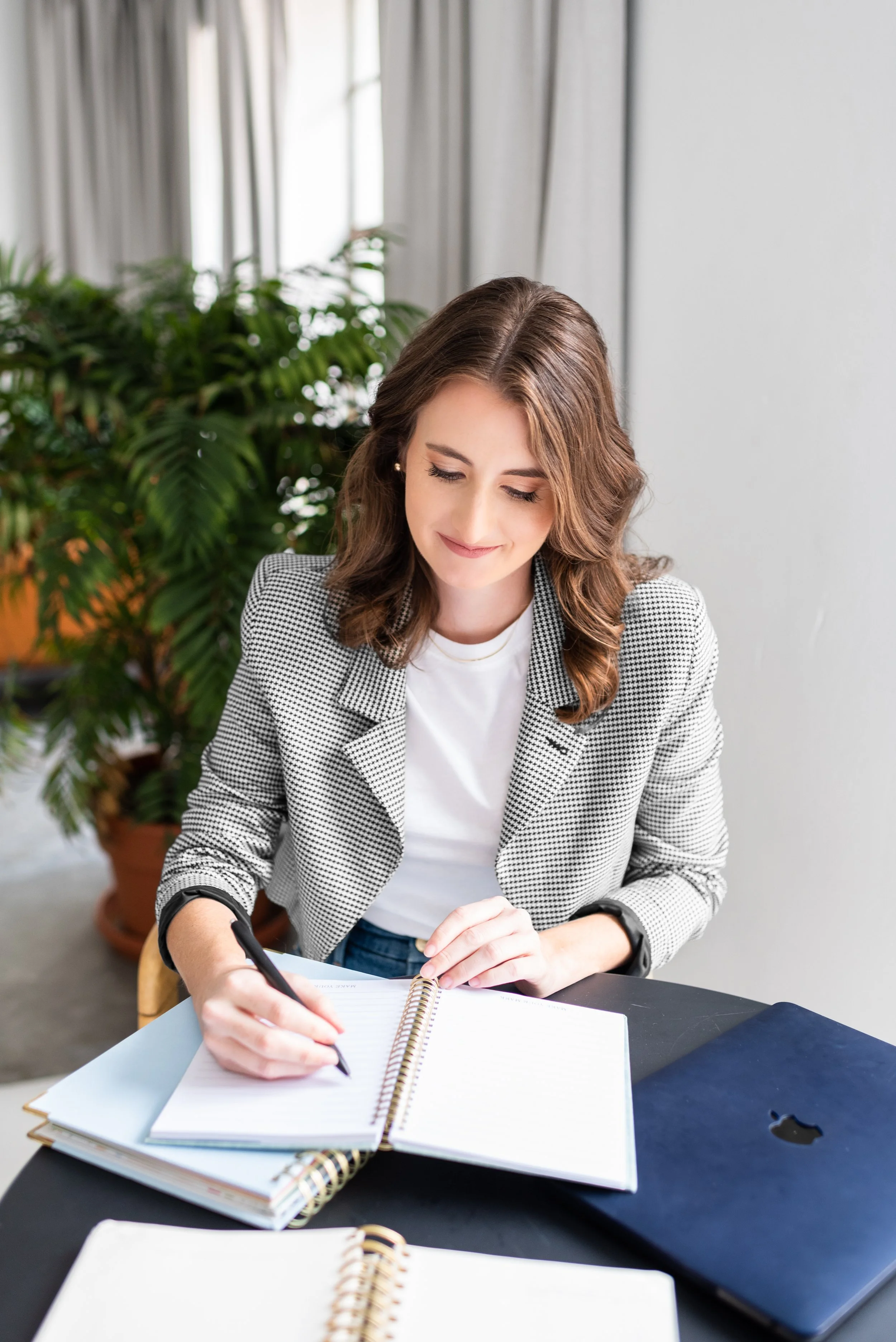 A woman with brown hair and a checkered blazer writing in a notebook at a desk with a closed laptop nearby, behind a large green potted plant.