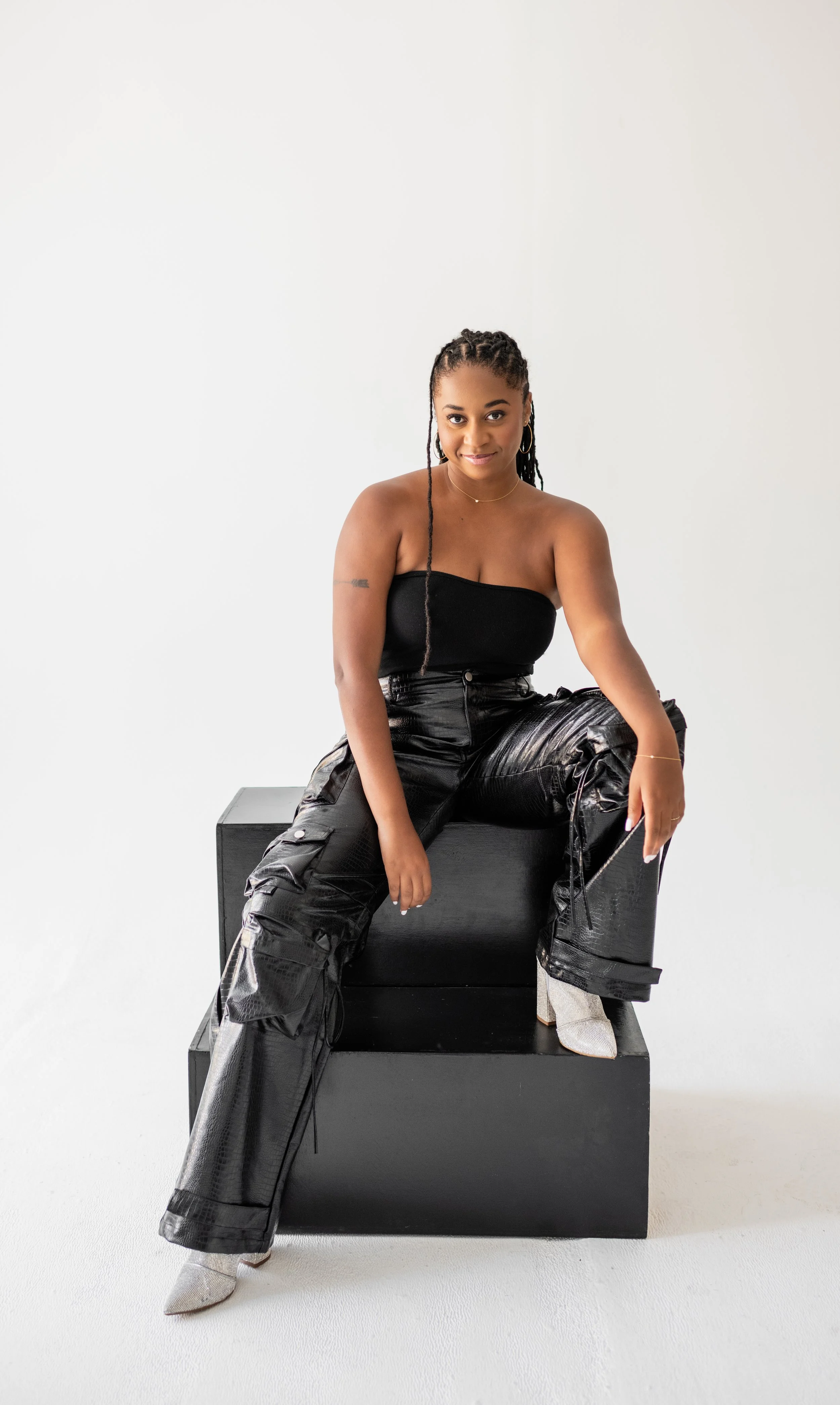 A woman with braided hair, wearing a strapless black top, shiny black cargo pants, and white high heels, sitting on a black block against a plain white background.