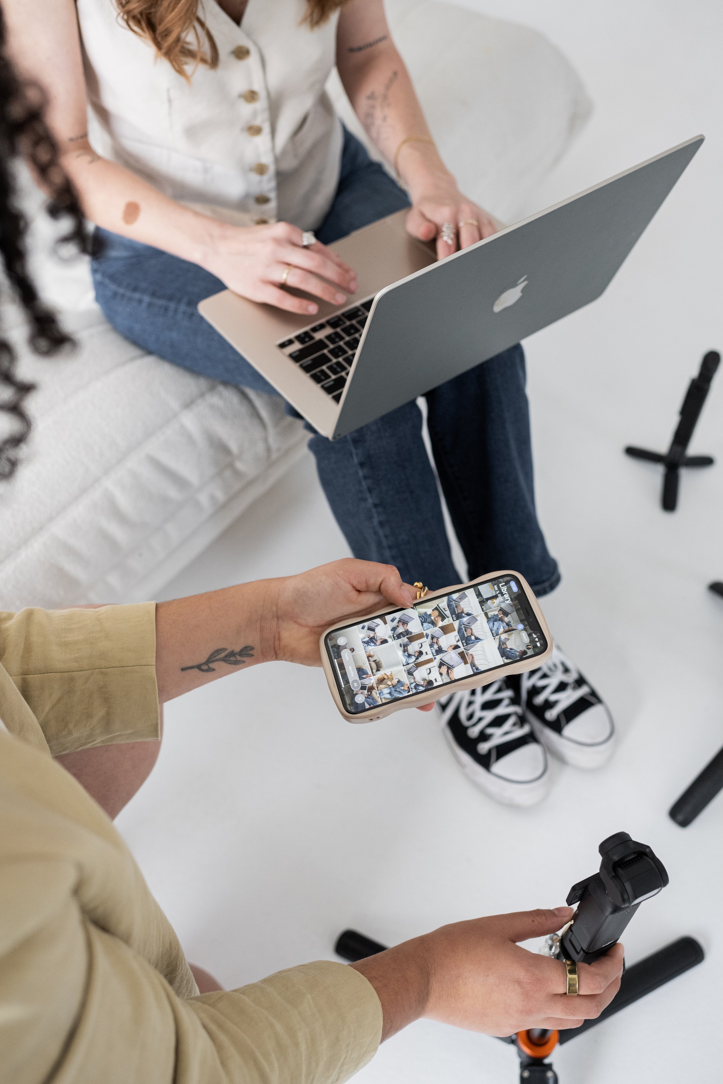 People preparing for a photoshoot or video recording, with one person holding a smartphone and a tripod, another working on a laptop, and a third standing nearby.