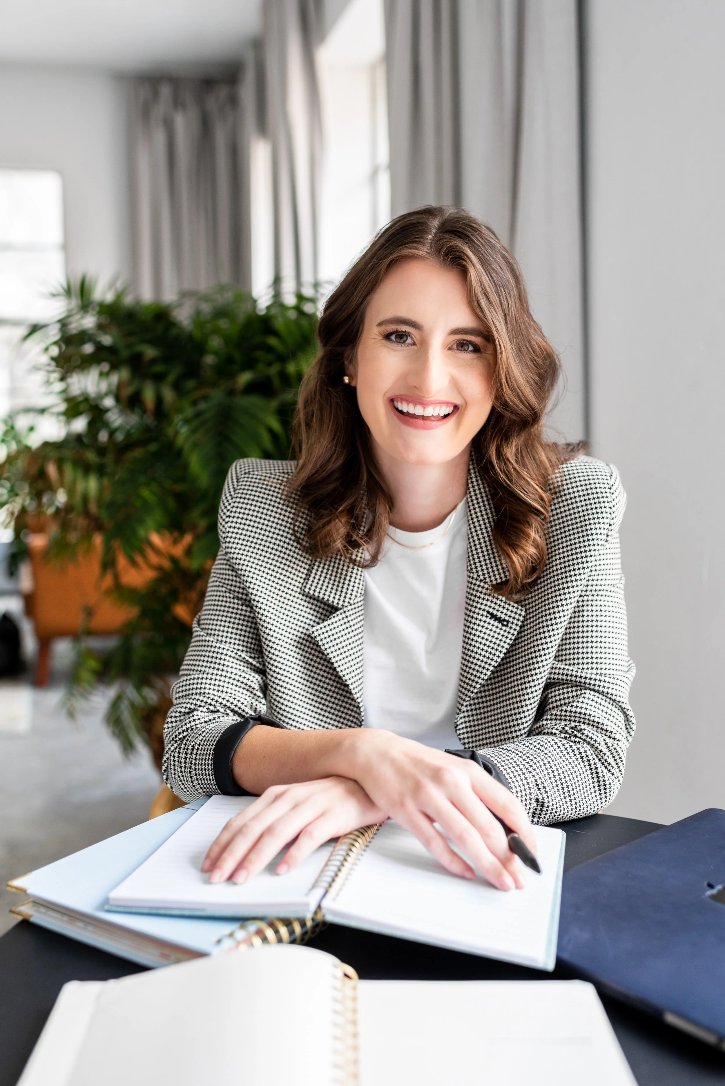 A young woman with shoulder-length brown hair, wearing a gray checkered blazer and white shirt, smiling and sitting at a desk with open notebooks, a pen, and a laptop in an office setting with large windows and green plants in the background.