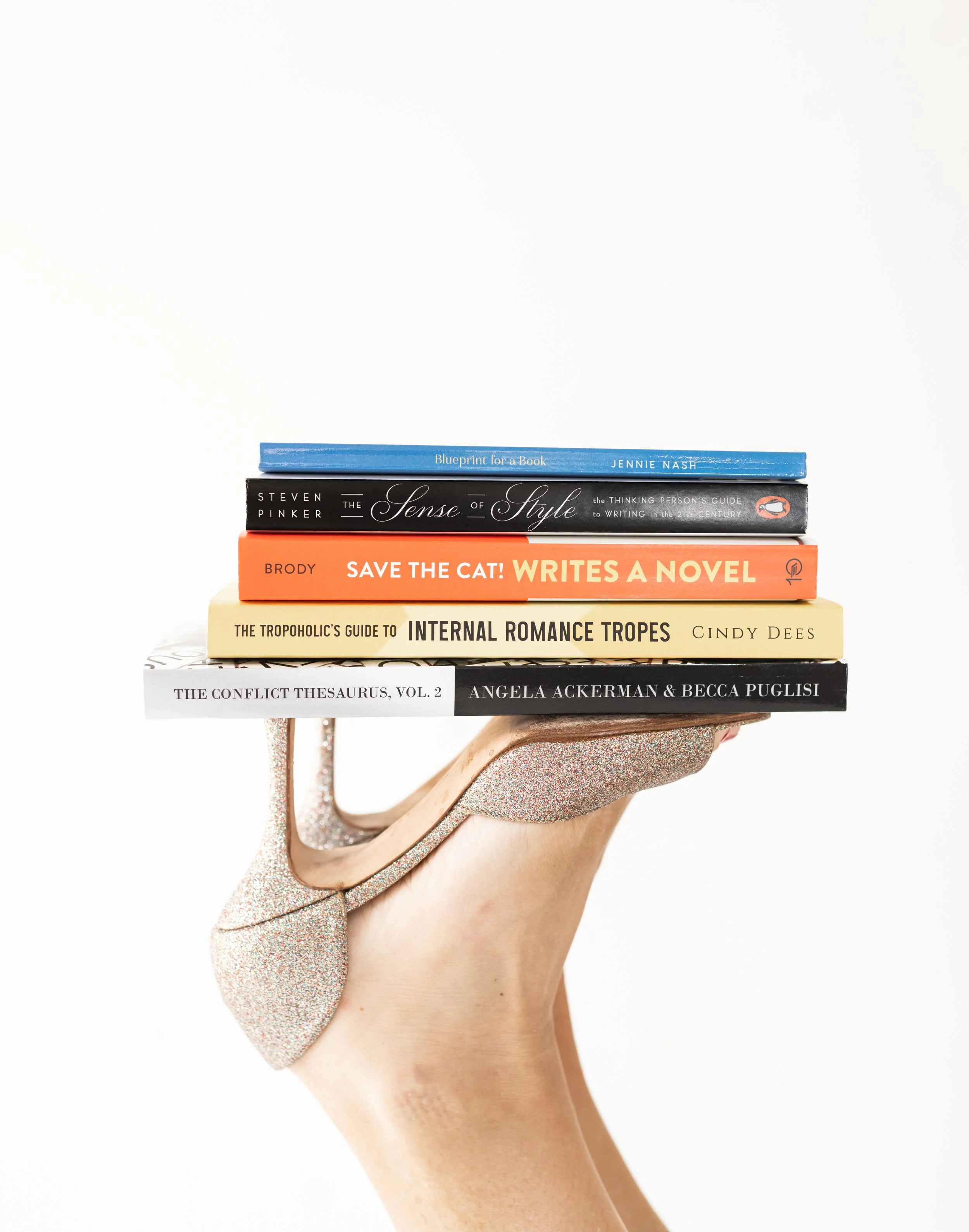 A hand wearing a sparkly high heel shoe holding a stack of six books on a plain white background.