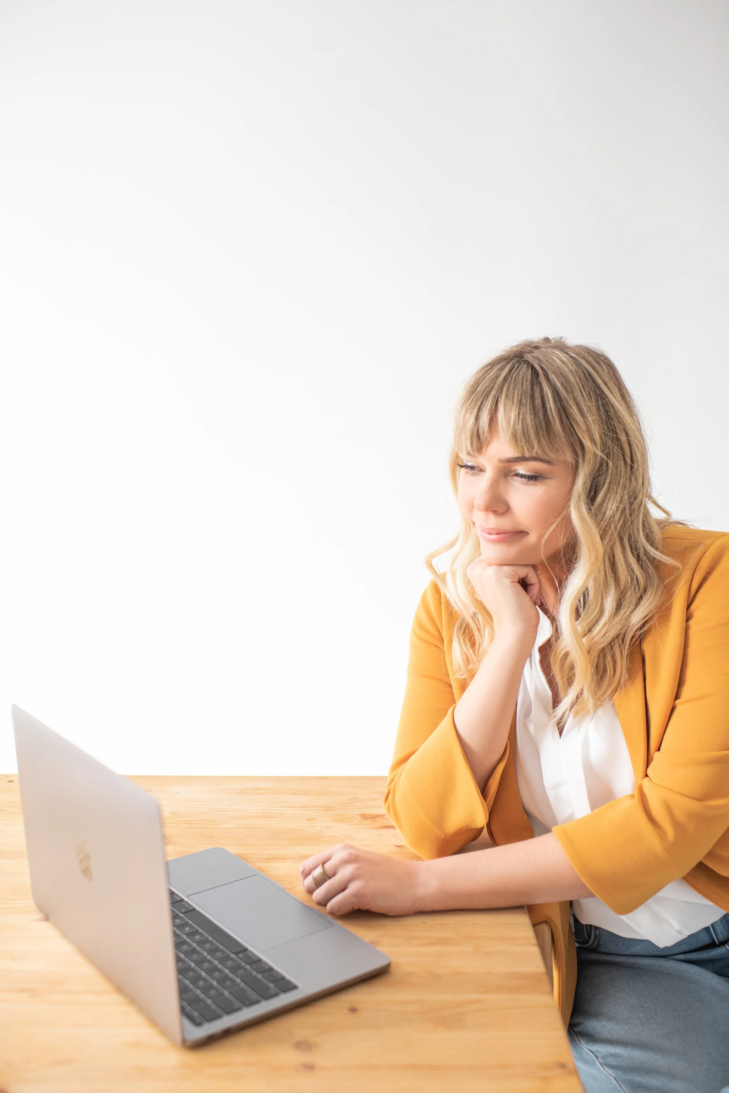 A woman with blonde hair, wearing a mustard yellow blazer and white shirt, sitting at a wooden table, looking at her laptop with a thoughtful expression.