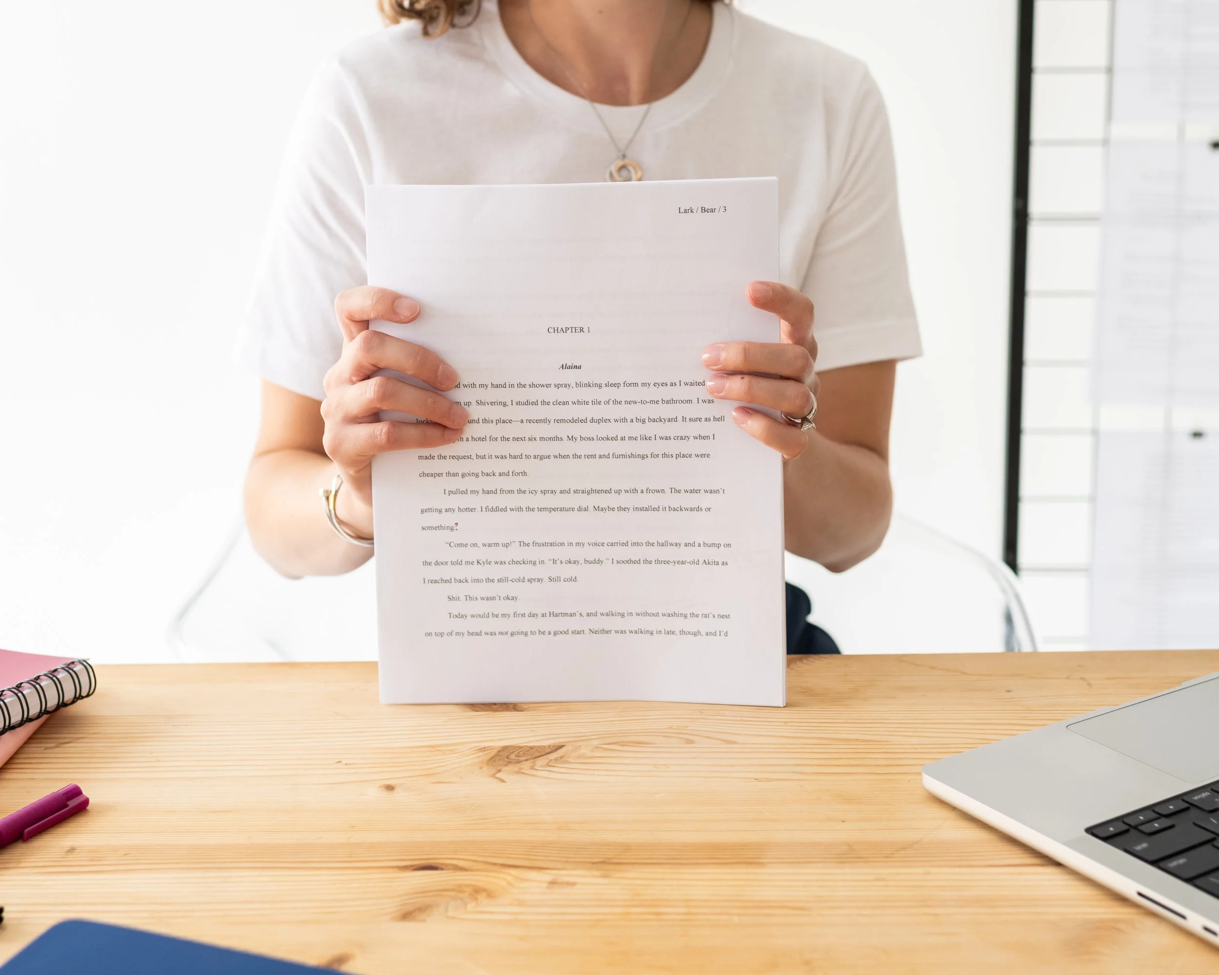 A woman in a white shirt holding a printed document at a wooden desk with a laptop, notebooks, and pens.