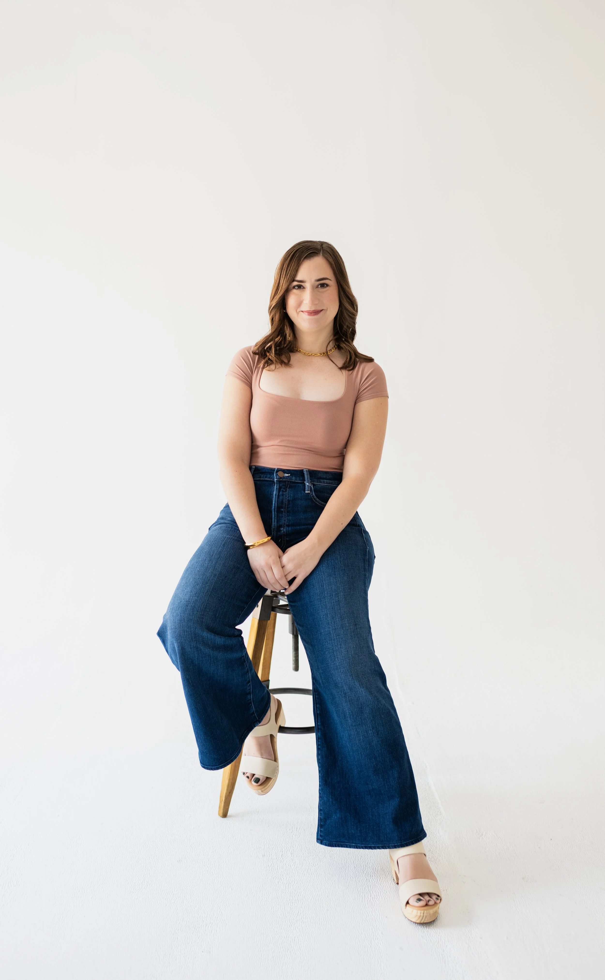 A woman with shoulder-length brown hair, wearing a pink top and blue jeans, sits on a stool in front of a white background, smiling at the camera.