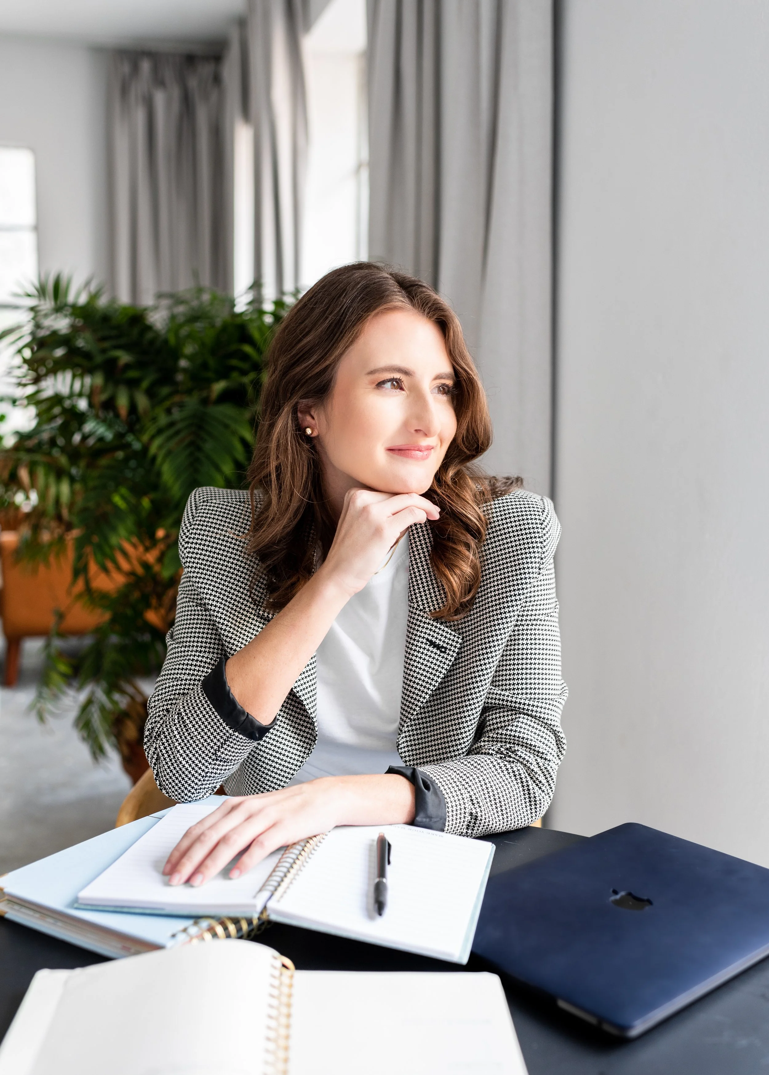 A woman sitting at a desk with a notebook, pen, and a laptop, looking thoughtfully out the window.