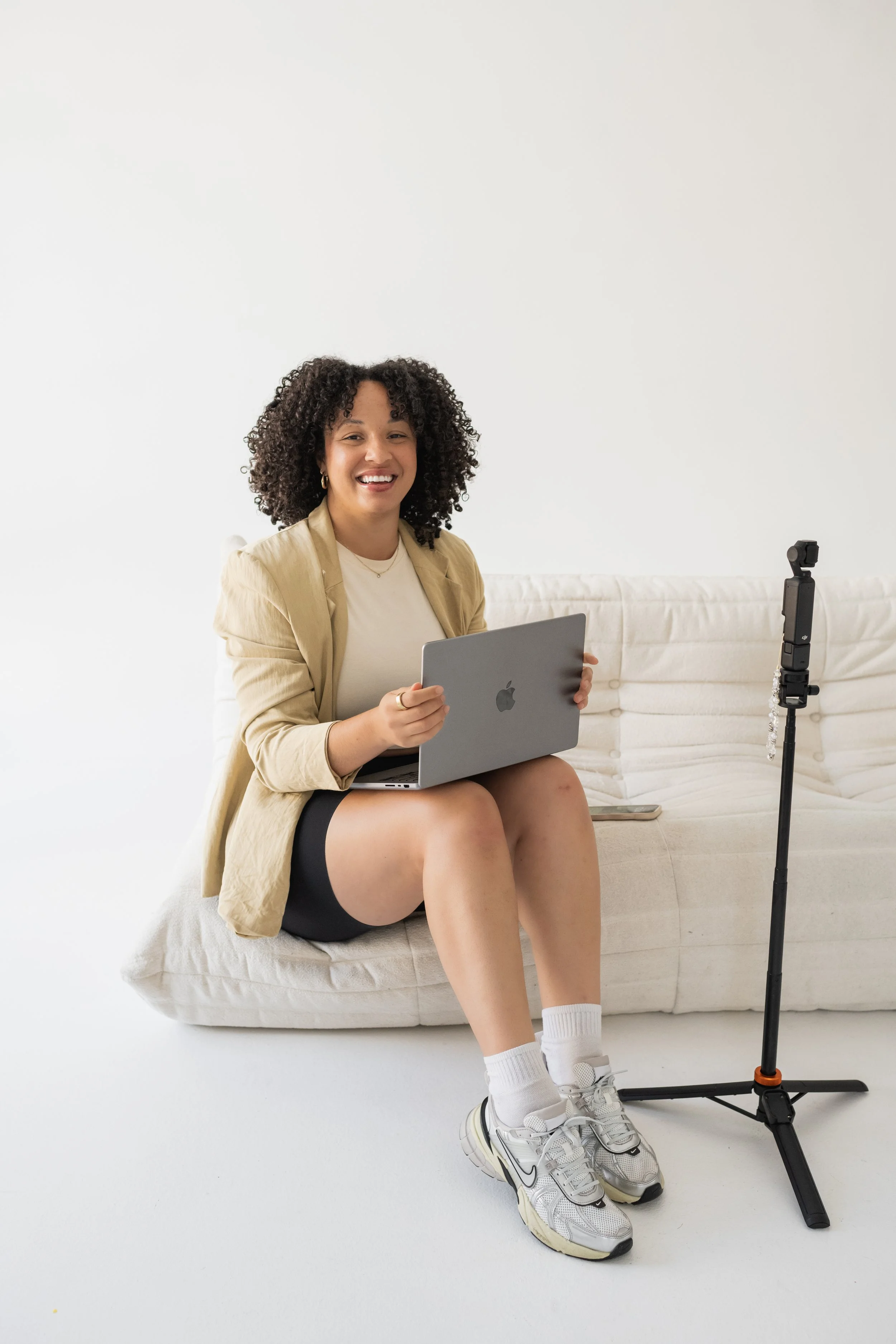 A woman sitting on a white couch, smiling, holding a laptop, with a phone and a tripod nearby.