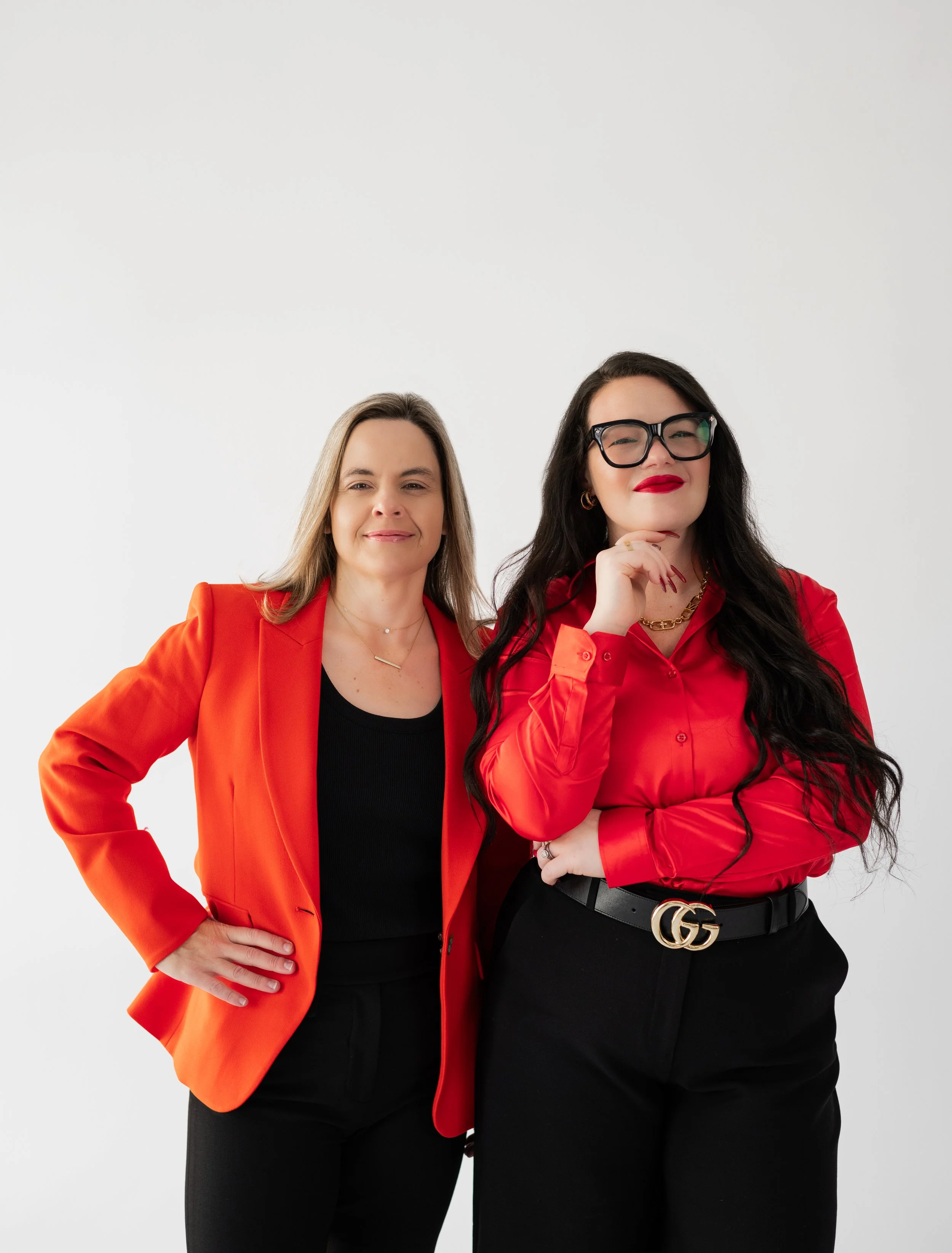 Two women standing together in stylish business attire against a plain white background. The woman on the left has shoulder-length light brown hair, wears an orange blazer and black shirt. The woman on the right has long dark hair, wears large black 