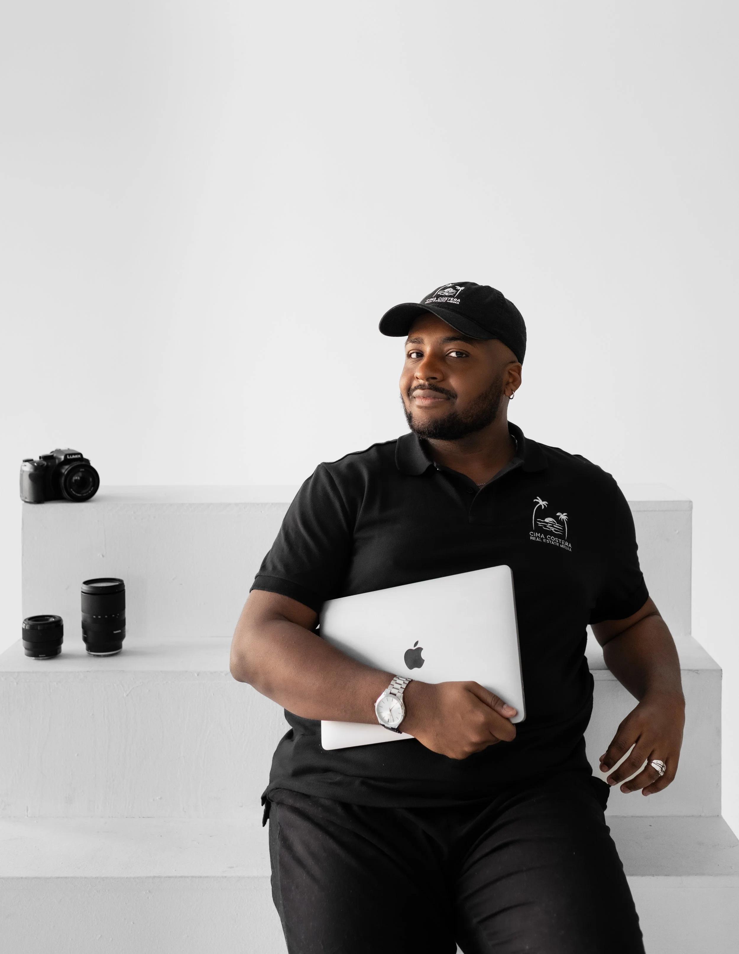 A man sitting and holding a silver MacBook, wearing a black polo shirt and cap, with cameras and lenses on a white shelf behind him.