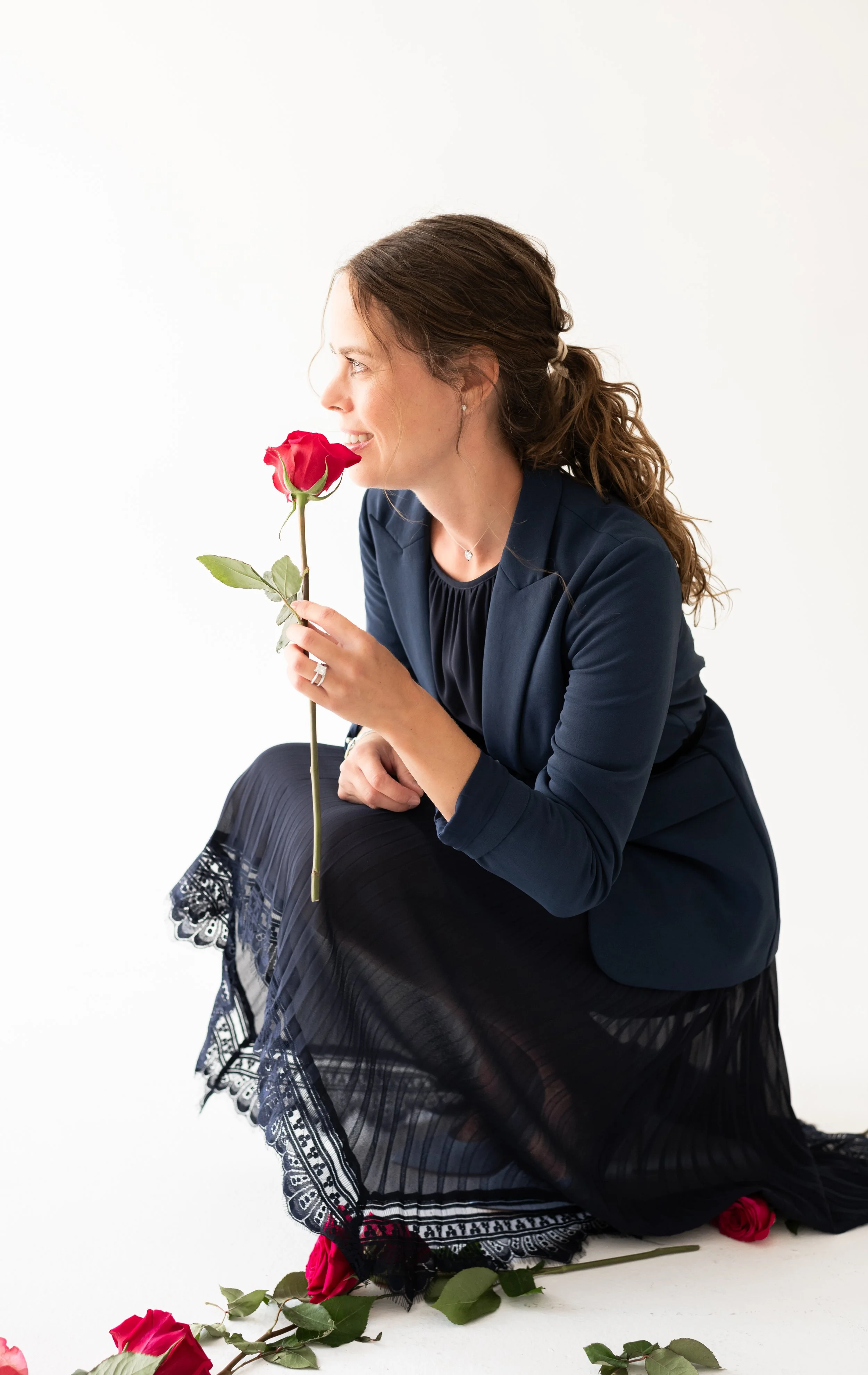 A woman with brown, curly hair styled in a ponytail, dressed in a navy blazer and a black dress with lace trim at the hem, kneeling on the floor and holding a red rose close to her face, with additional roses scattered around her on a white backgroun