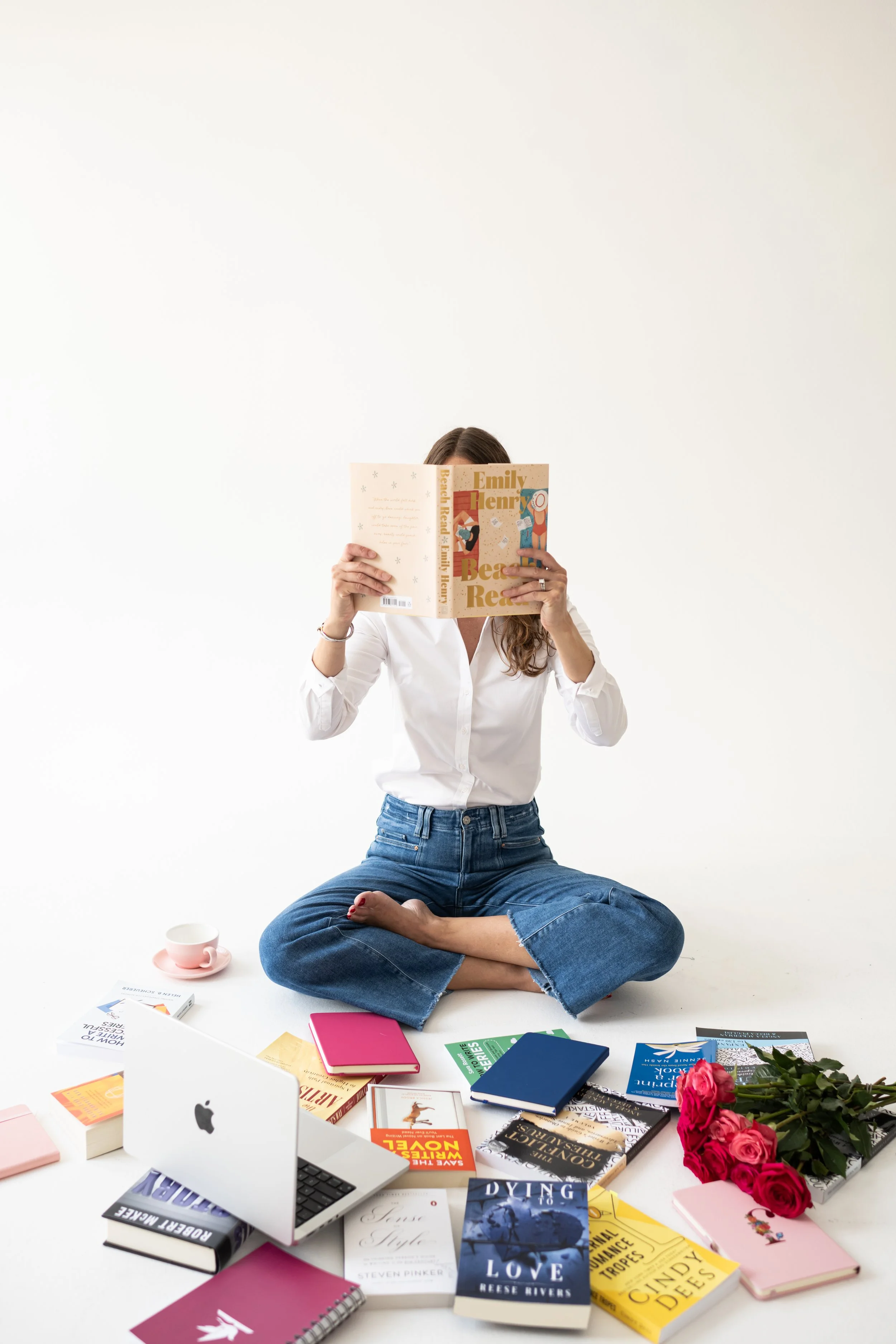 A woman sitting cross-legged on the floor reading a book with a blank white wall in the background. Surrounding her are multiple books, a laptop, pink and blue notebooks, a pink cup and saucer, a bouquet of pink roses, and a few other items.