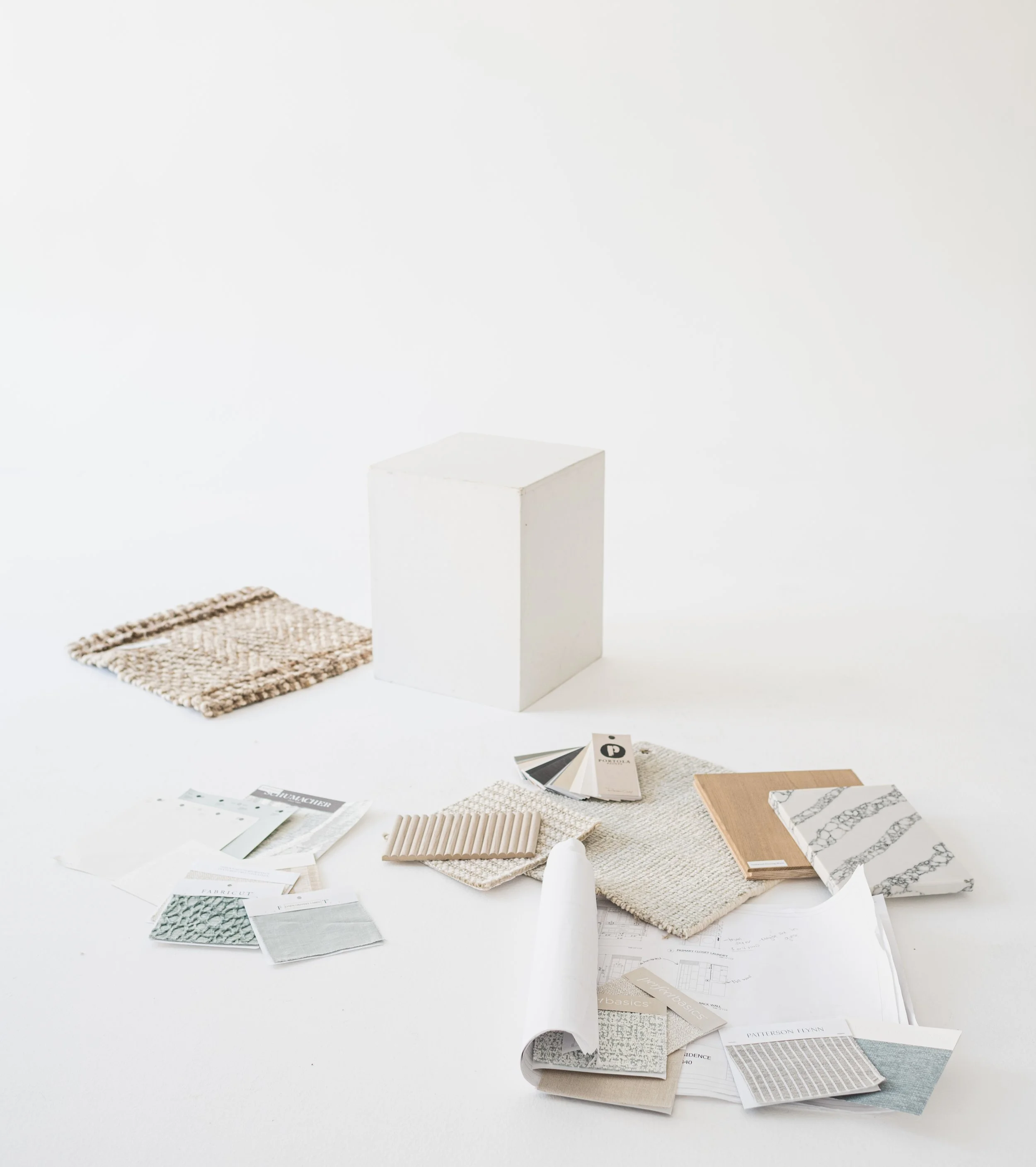 Design or interior decorating samples including fabric swatches, paint color cards, and material samples on a white surface with a blank white cube and a woven fabric piece.