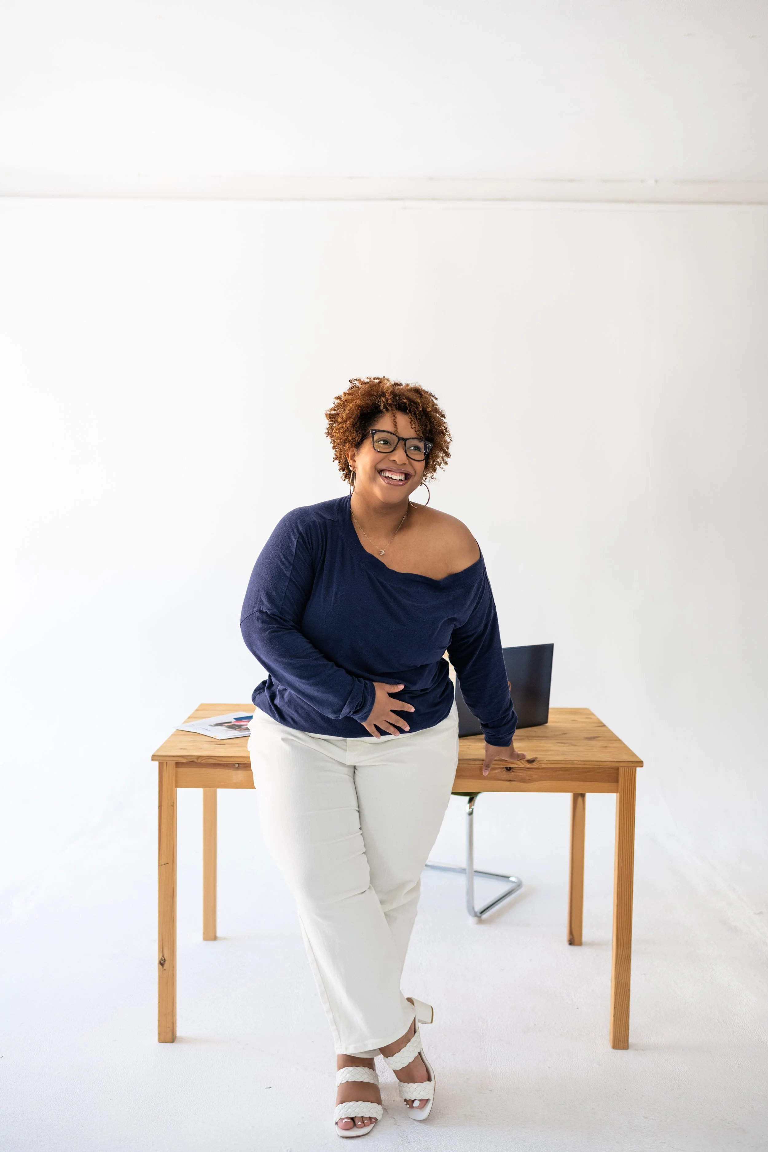 A smiling woman with curly hair and glasses leaning against a wooden desk in a bright, minimalistic office space.