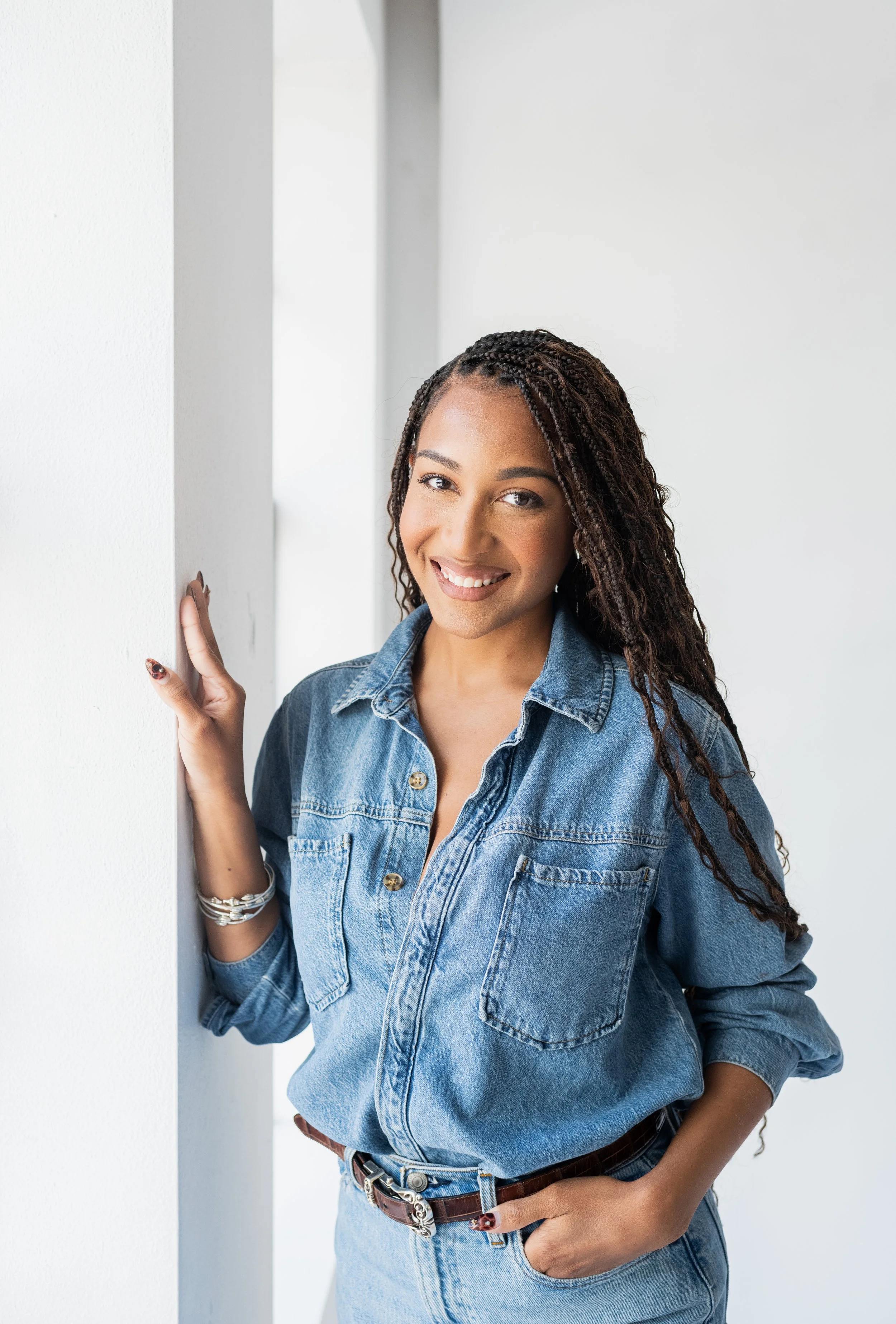 Woman with long curly hair, smiling, wearing a denim shirt and jeans, standing by a white wall.