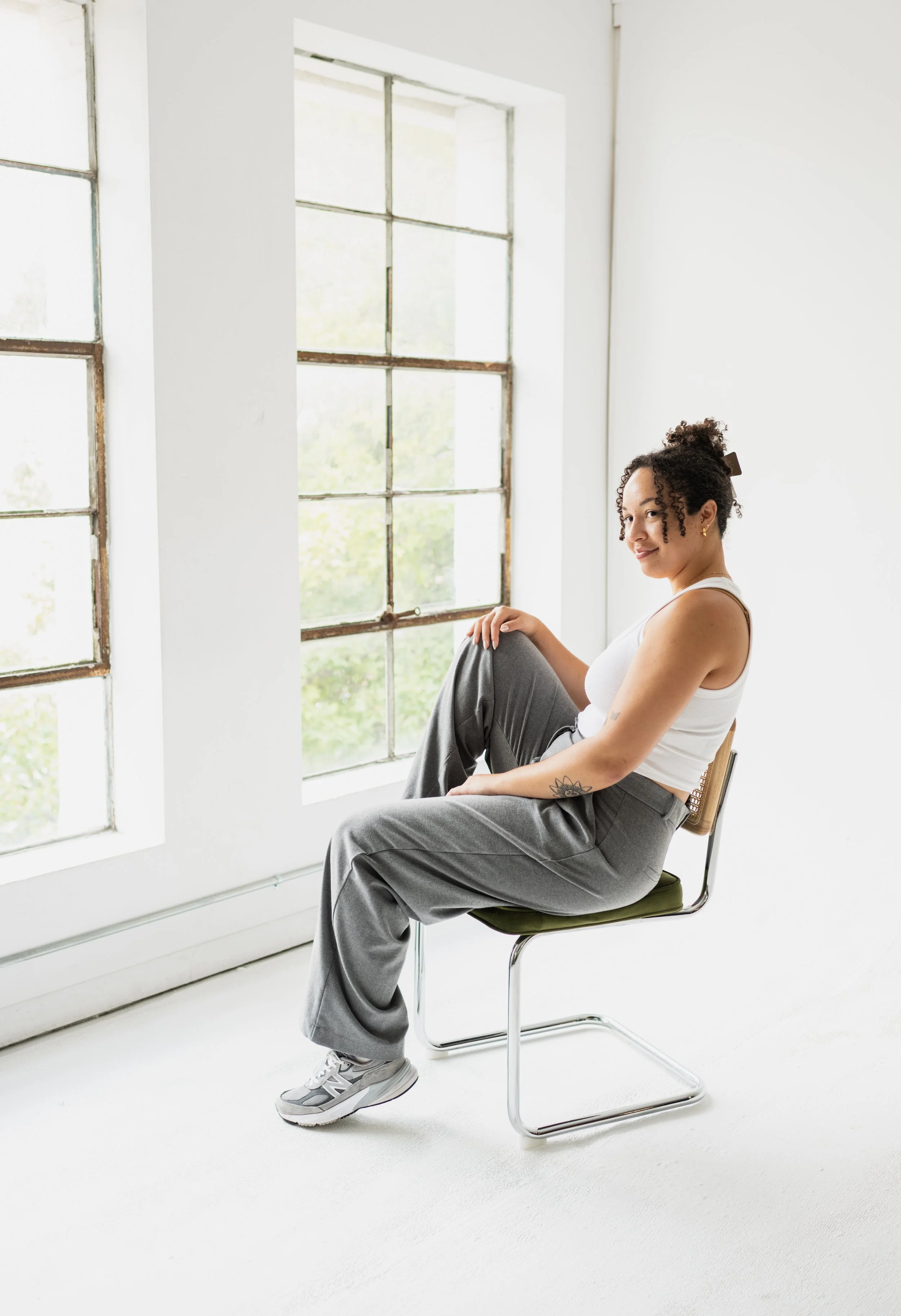 A woman sitting on a green chair near a large window with a white wall background, wearing a white tank top, gray pants, and sneakers, smiling at the camera.