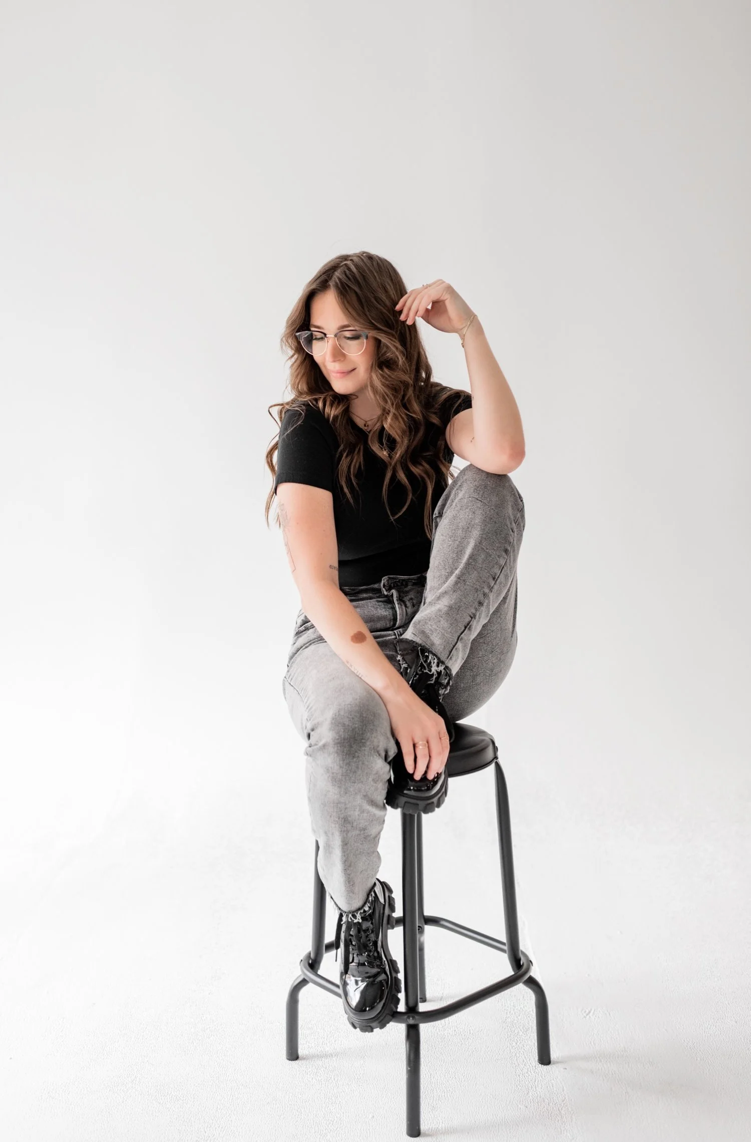 A woman with wavy brown hair, wearing glasses, a black t-shirt, gray jeans, and black boots, sitting on a black stool in a plain white studio background, looking down and smiling.